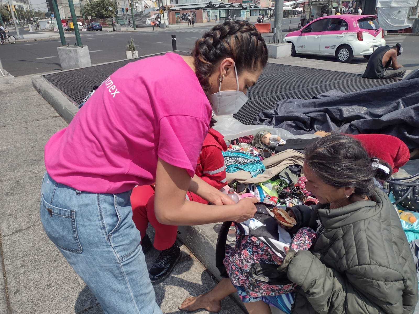 Una mujer con mascarilla y peinado trenzado ayuda a una mujer adulta que está sentada en el suelo con una cobija, en un mercado callejero con ropa y objetos dispersos. En el fondo, hay autos y una calle con árboles y personas.
