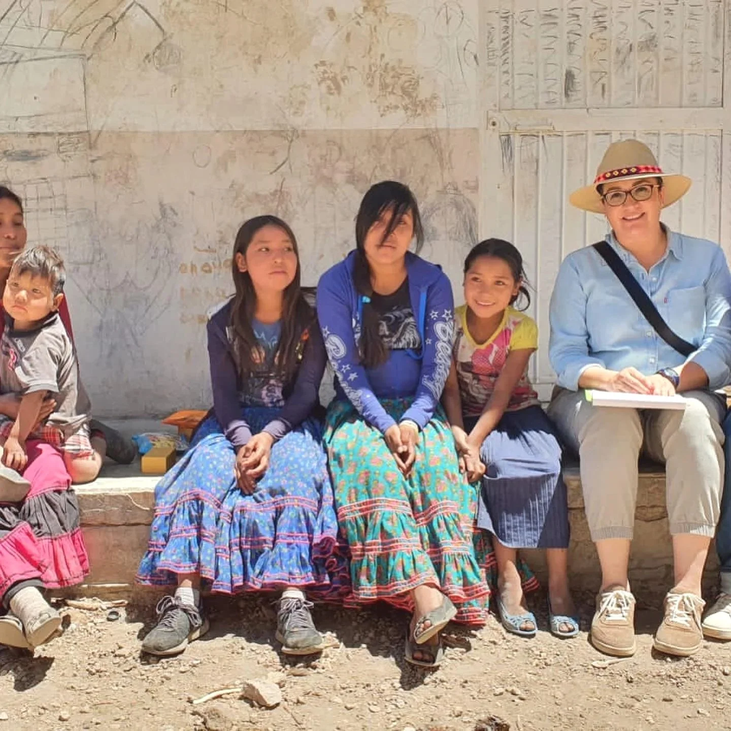 Grupo de personas sentadas en una banca, con una pared de fondo. Incluye tres niñas con ropa colorida y un adulto con sombrero y gafas, todos sonriendo o con expresión tranquila.