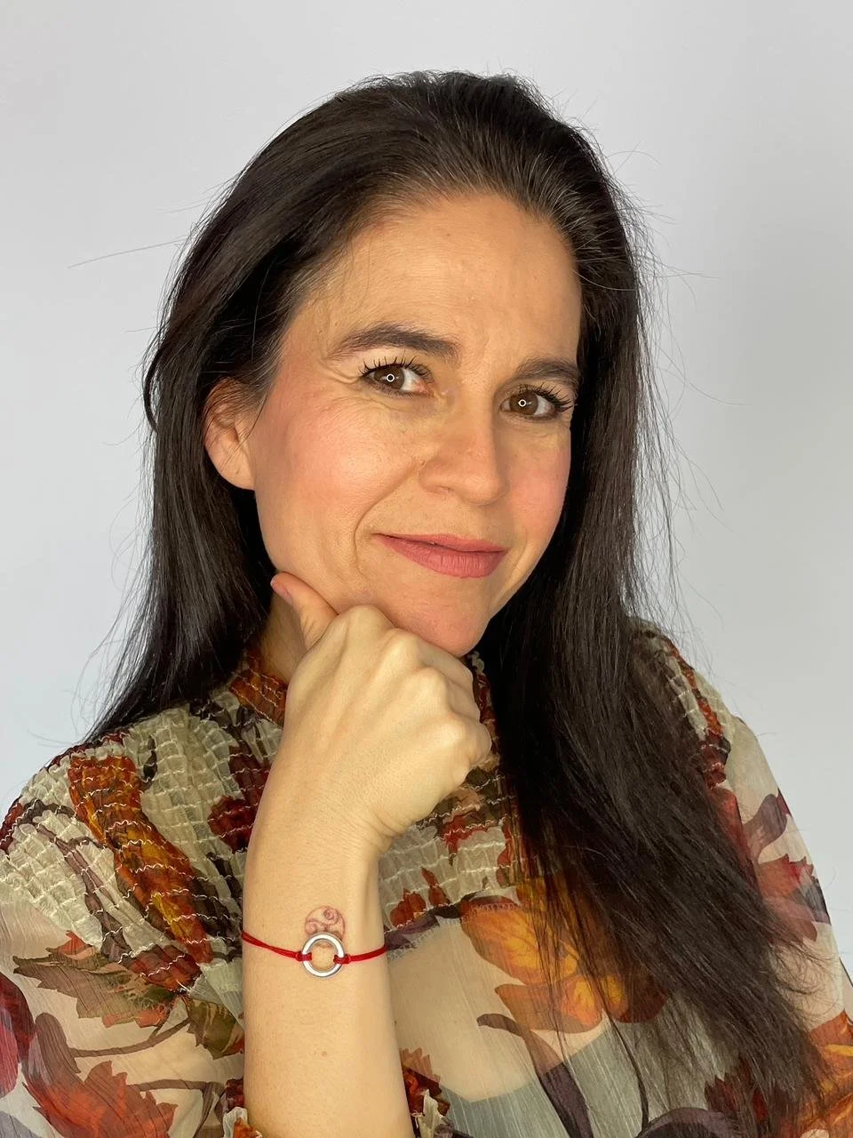 Retrato de una mujer con cabello castaño oscuro y expresión pensativa, usando un vestido con estampado de hojas y un pulsera roja con un símbolo circular en la muñeca.