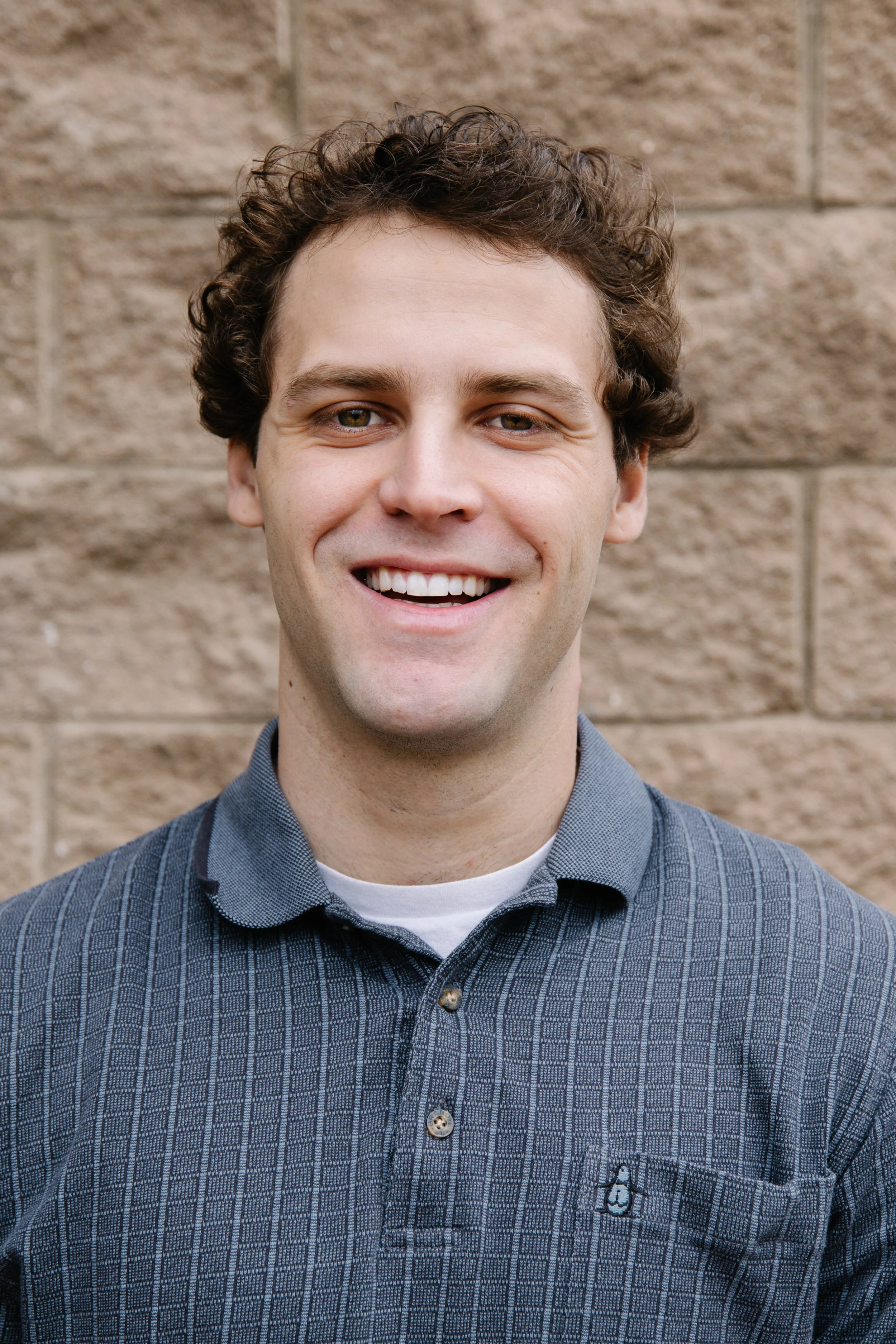A young man with curly brown hair smiling in front of a brick wall, wearing a blue polo shirt with a small penguin logo.