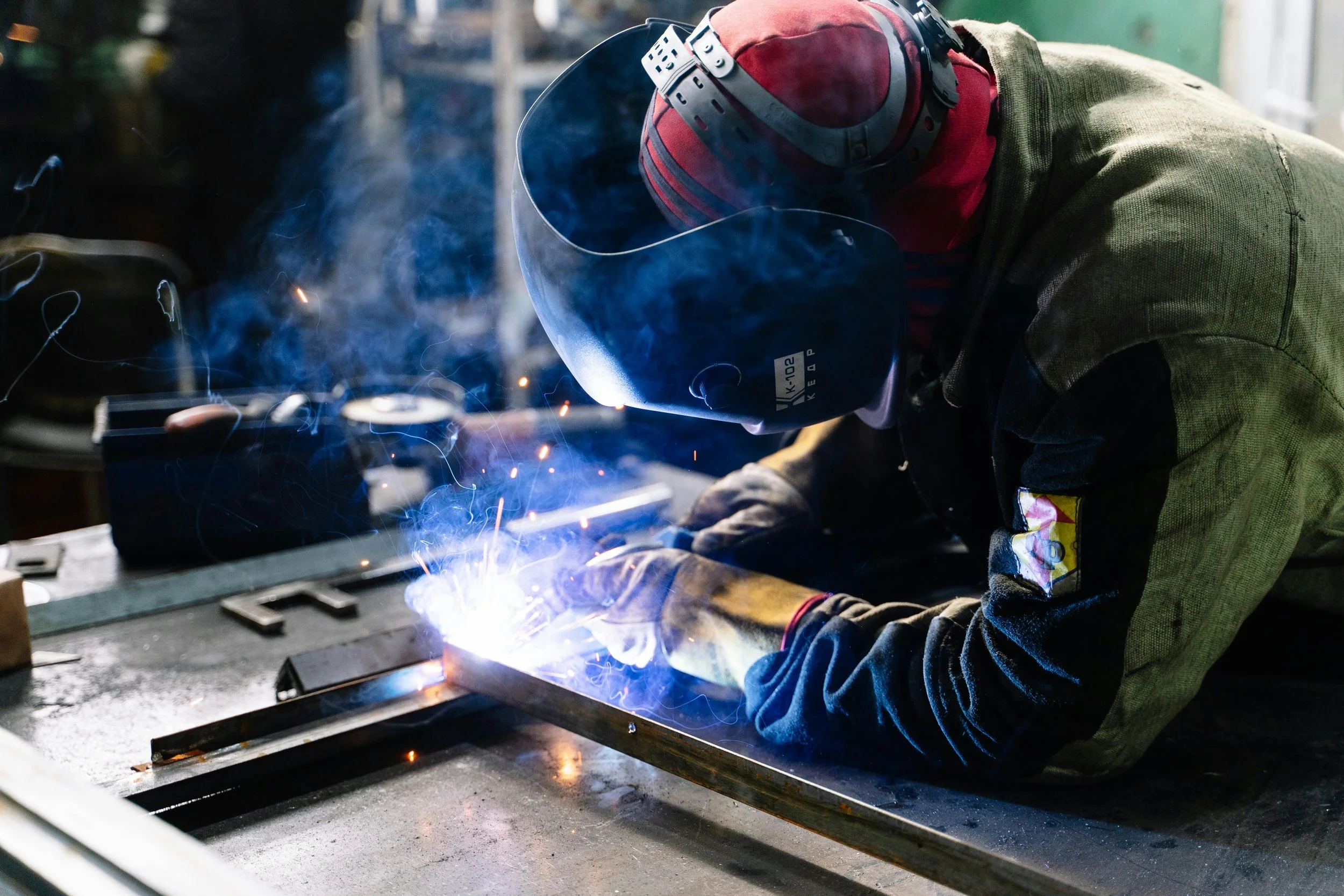 A welder wearing a helmet and gloves welding metal on a workbench in a workshop, with sparks and blue light.