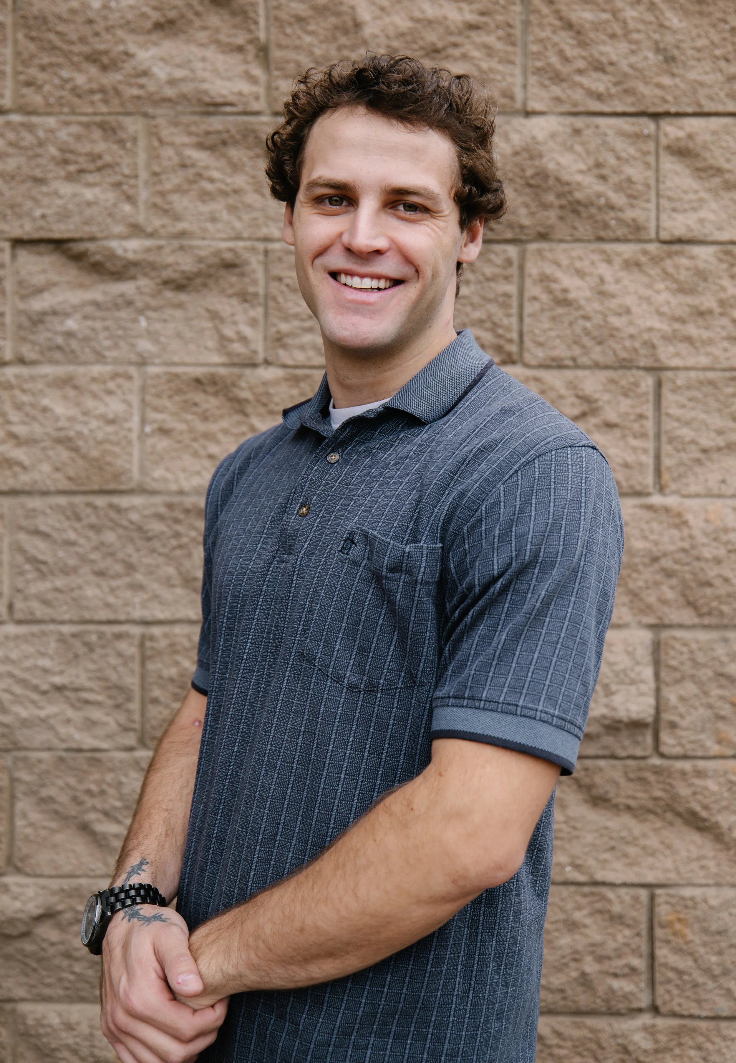 A young man with curly brown hair smiling and standing in front of a brick wall, wearing a gray patterned polo shirt, a watch, and a bracelet.