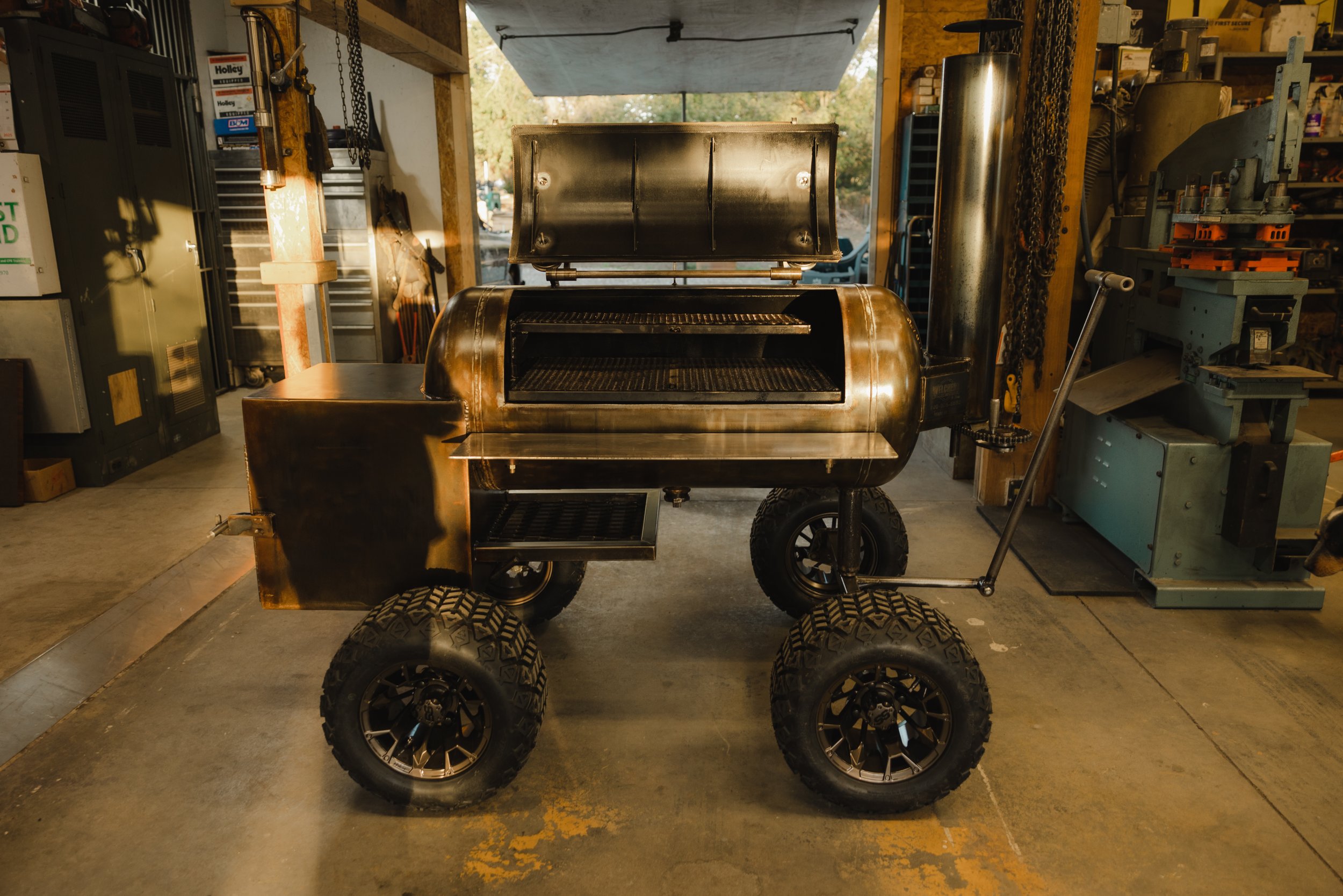 A metallic mobile grill with large tires, set in a workshop. The grill has an open lid showing cooking grates inside.