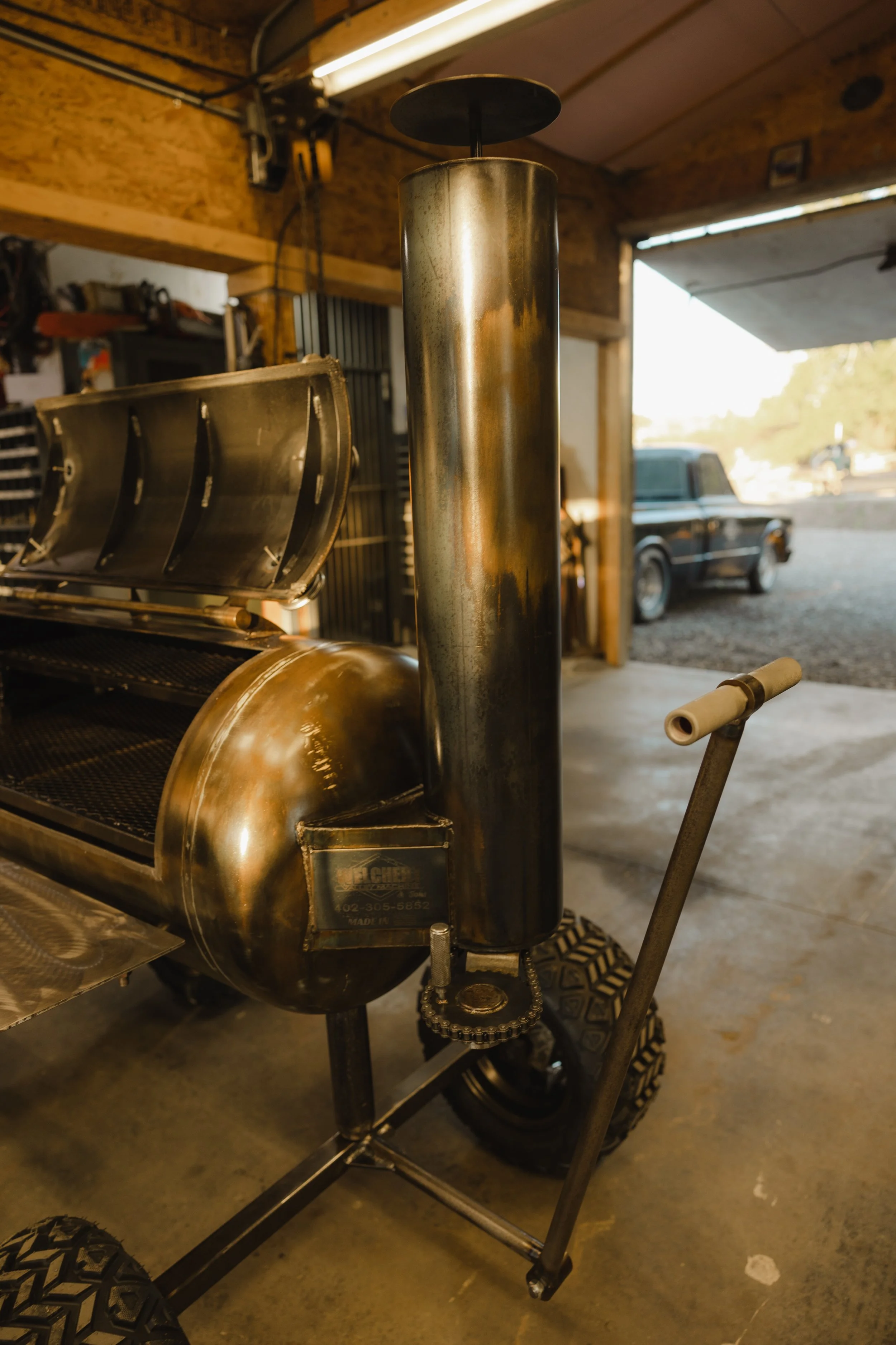 Close-up of a metallic outdoor smoker grill with a vertical chimney and a side handle, in a garage with a view of a parking lot and vintage truck outside.