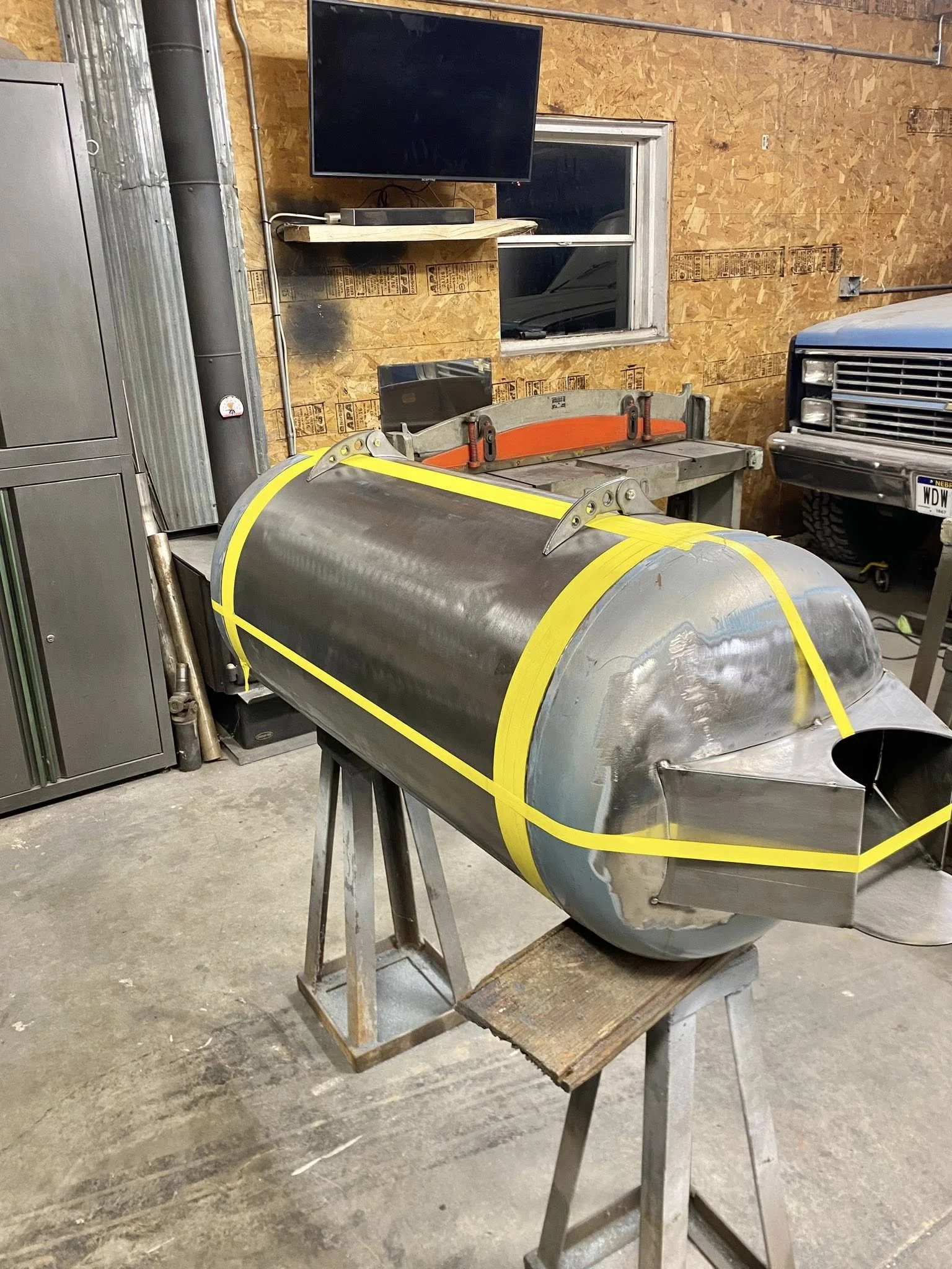 A metal pipe with yellow tape on it, mounted on a stand in a workshop with tools, a television, a window, and a vehicle in the background.