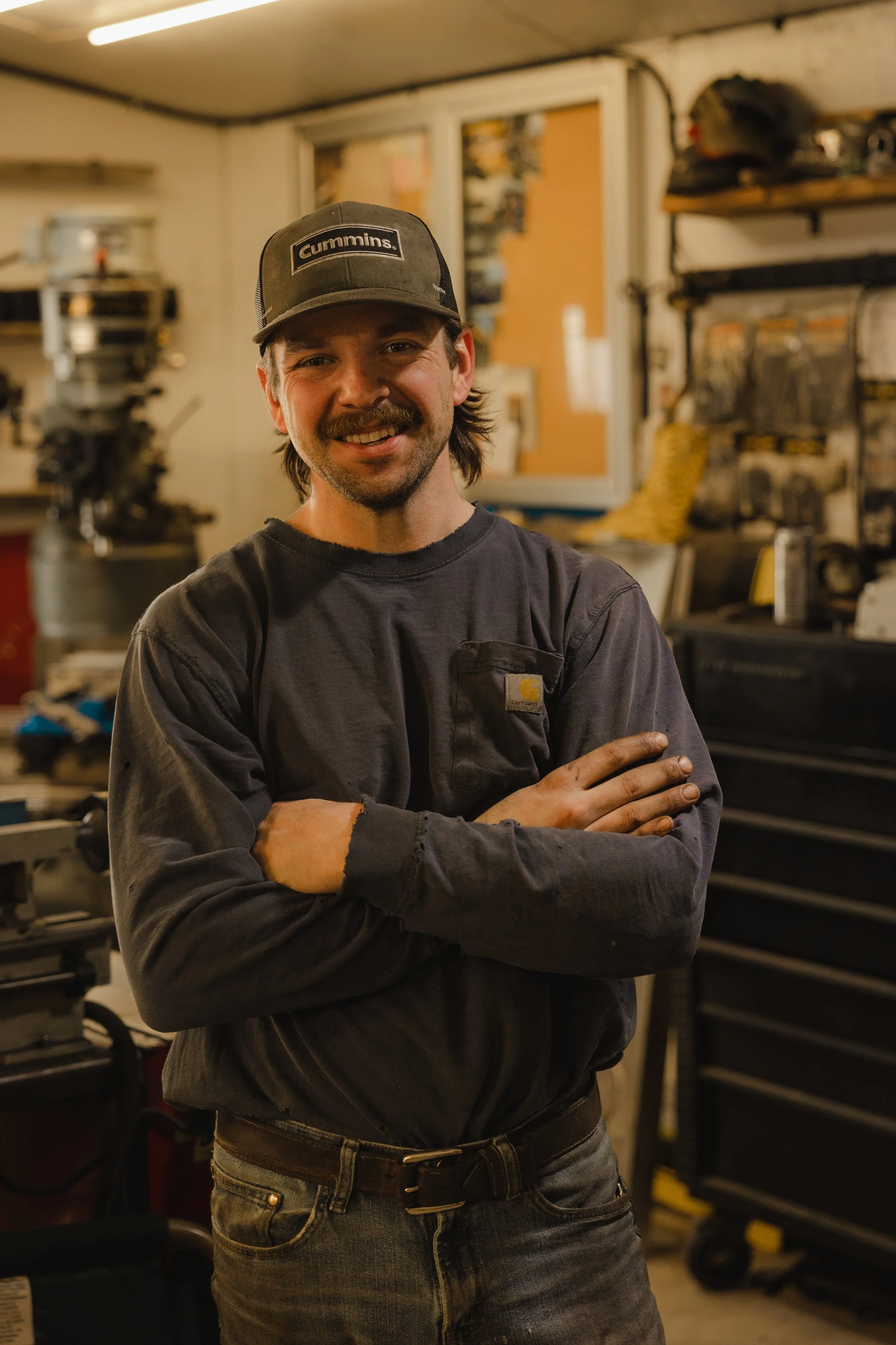A young man with a beard and mustache in a workshop, wearing a dark gray Carhartt shirt and a cap with the Cummins logo, standing in front of a pegboard with tools and a yellow toolbox, smiling with arms crossed.