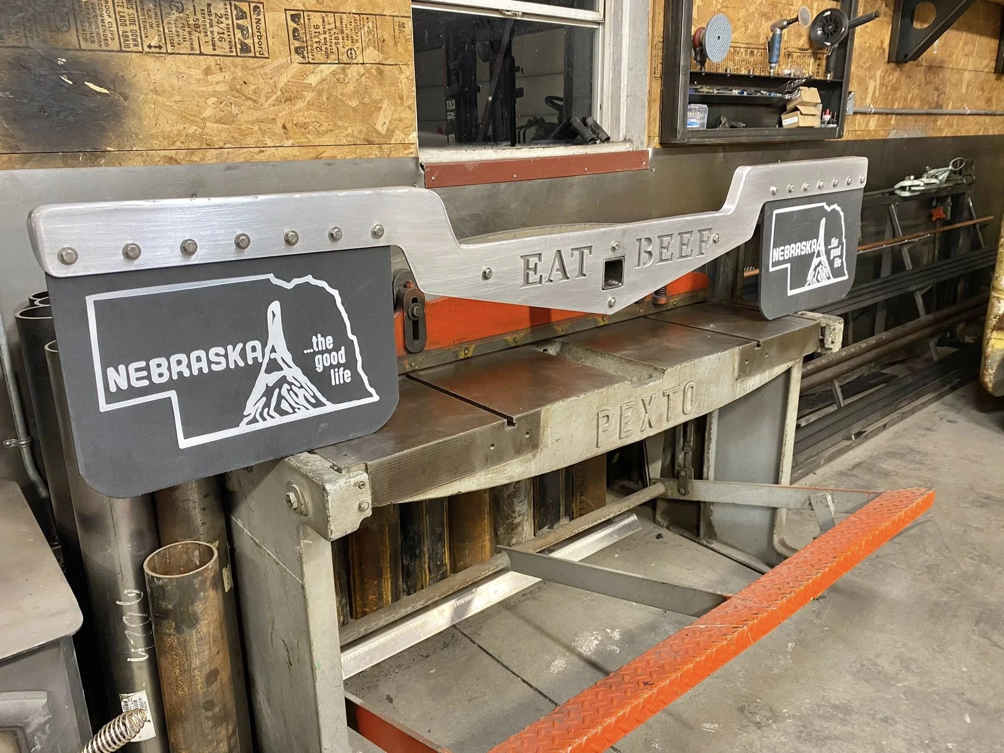 Metal sign with cutout text that reads 'EAT BEEF' on a workbench in a workshop. There are two Nebraska-themed stickers on the sign, featuring the outline of Nebraska and a mountain scene with the slogan 'the good life'.