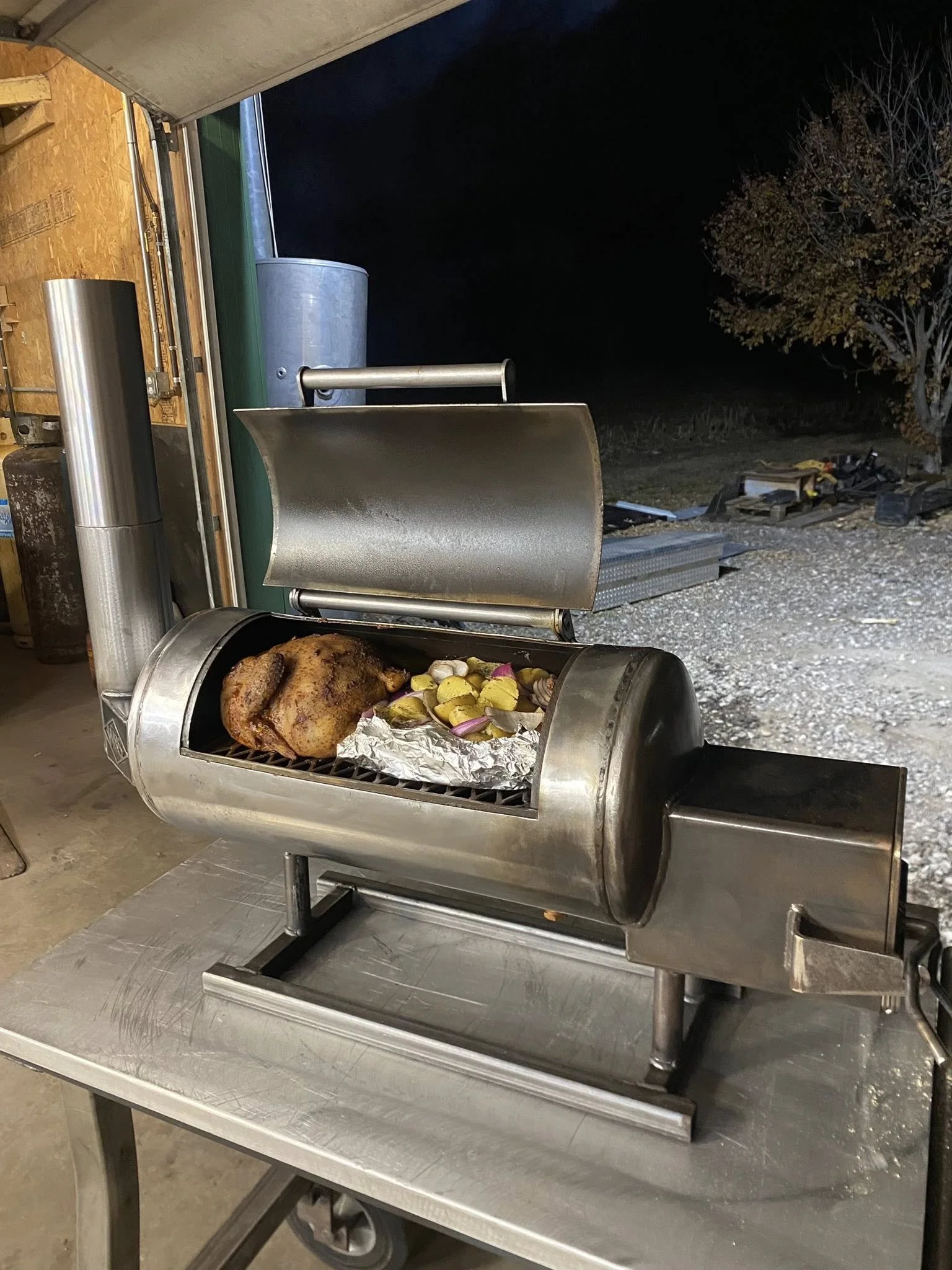 A stainless steel meat grinder with a roasted whole chicken and cut vegetables inside, placed on a metal table in a garage or workshop at night.