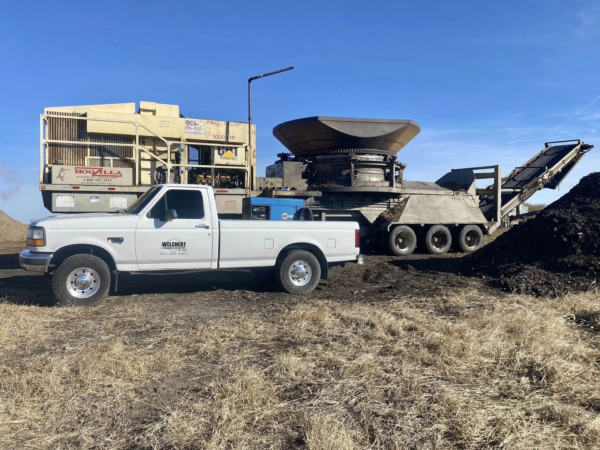 Construction site with a large industrial machine, a white pickup truck with company logo, and piles of dirt under a clear blue sky.