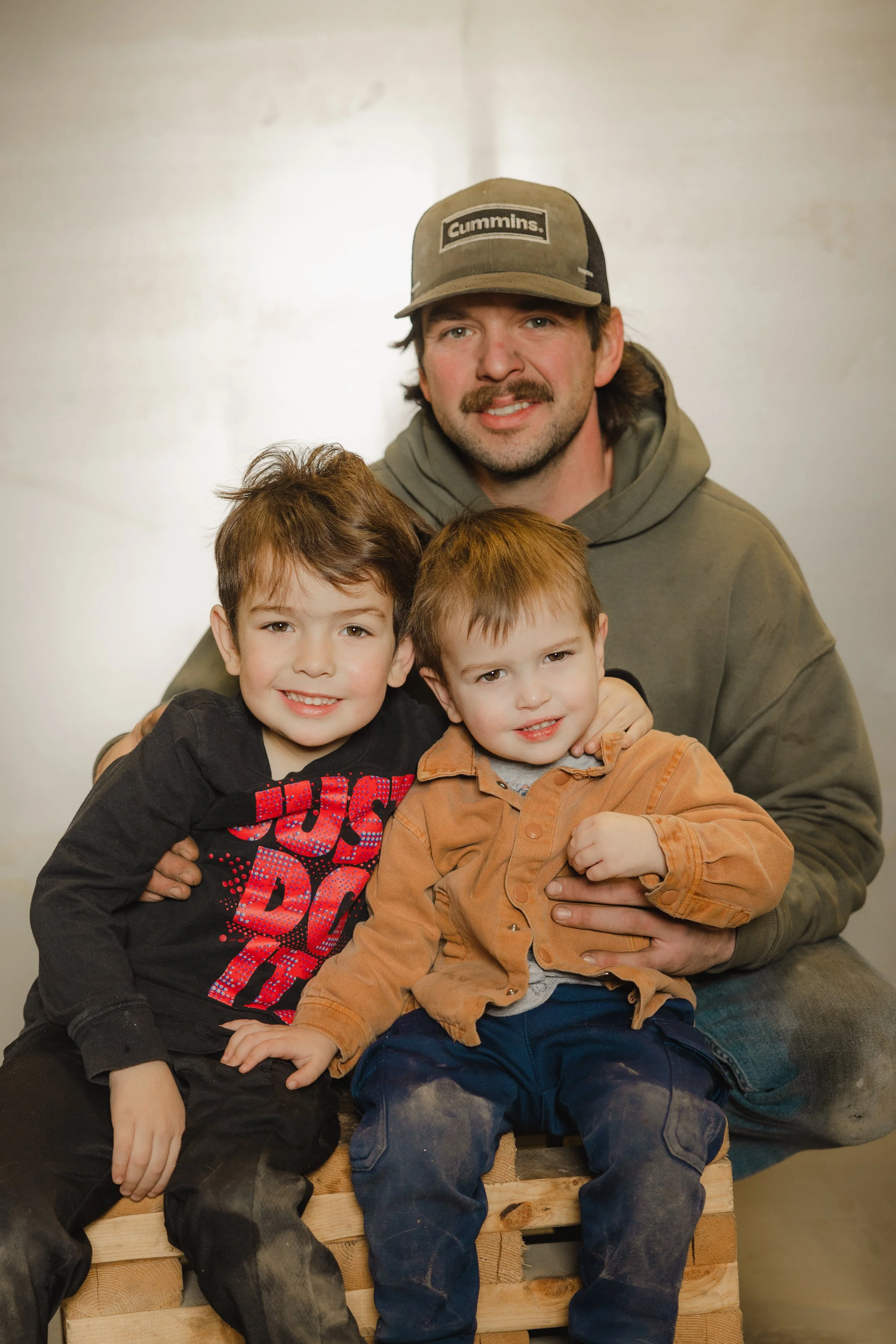 A man with two young boys seated on a wooden crate, smiling at the camera. The man is wearing a baseball cap with 'Cummins' logo and a hoodie, while the boys are dressed in casual clothes with one in a black shirt and the other in an orange shirt, both showing signs of dirt and work.