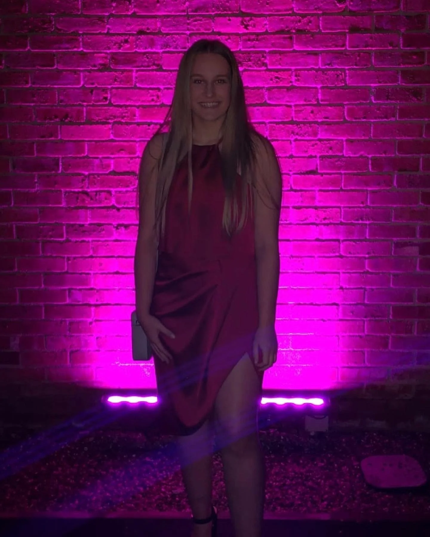 A woman in a red dress standing against a pink brick wall illuminated by pink and purple lighting.