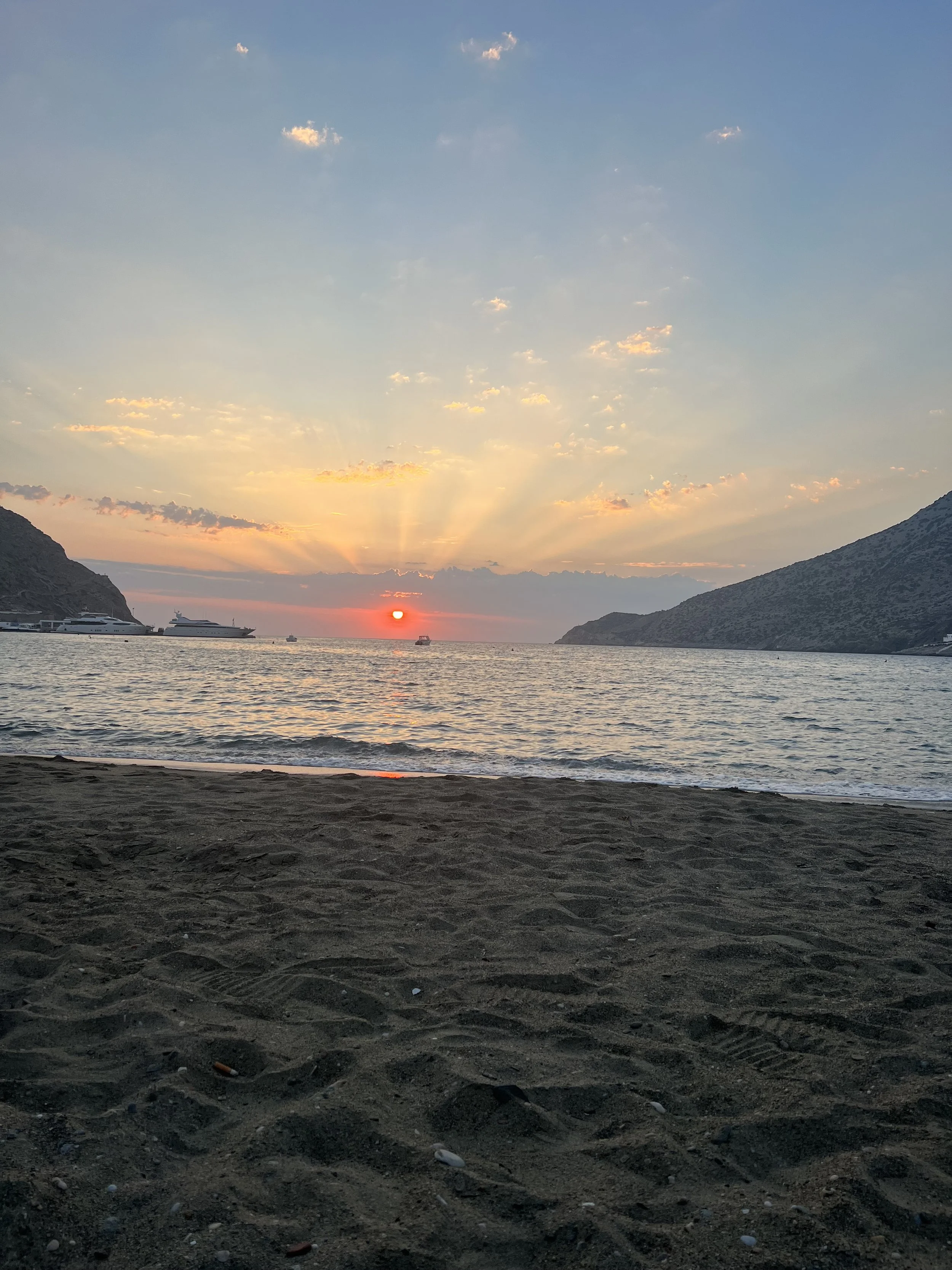 Sunset over the ocean with mountains on both sides, boats in the water, and sandy beach in the foreground.