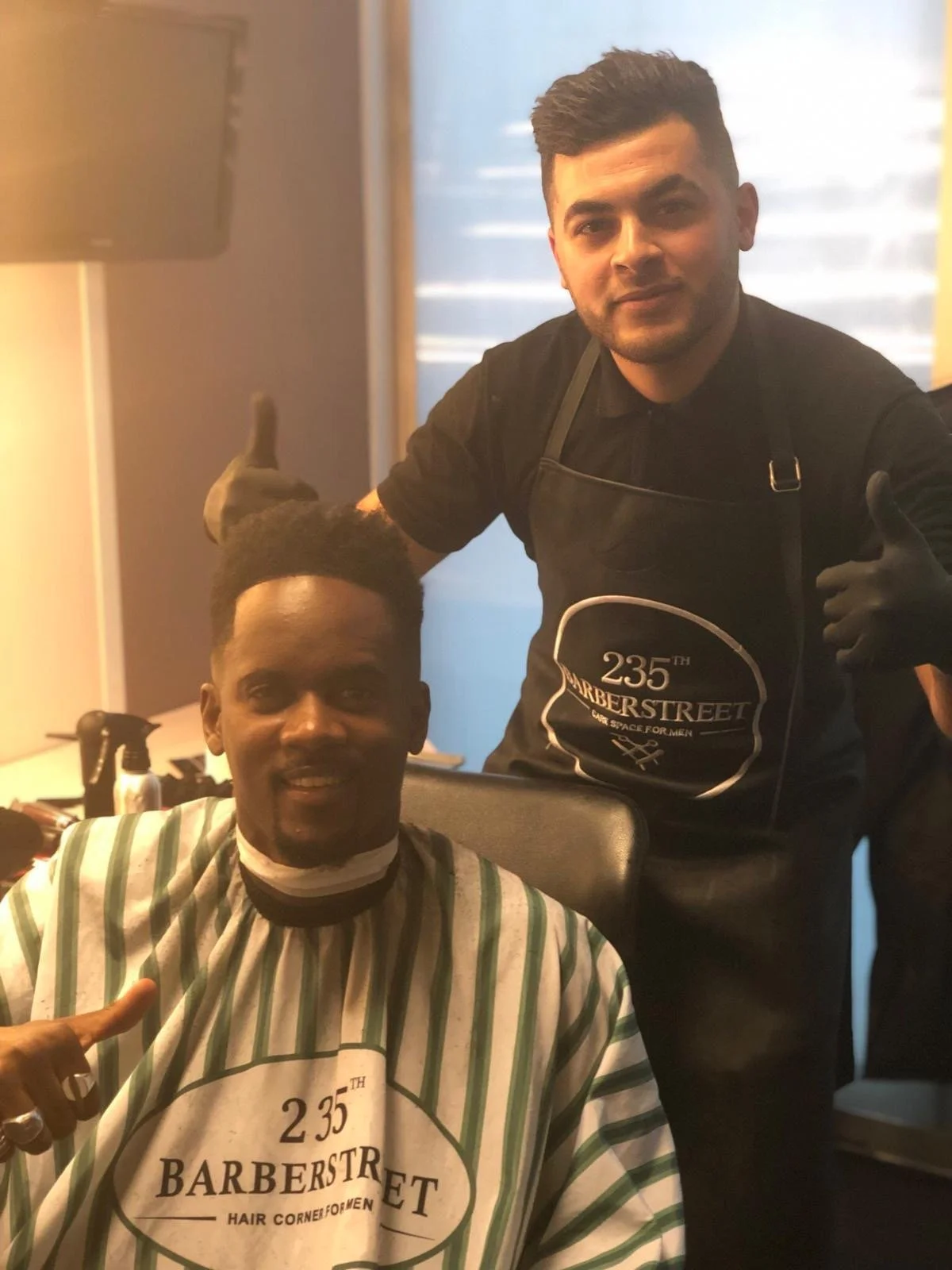 Deux hommes dans un salon de coiffure, un homme reçoit une coupe de cheveux et porte une cape de barber. L'autre homme, un coiffeur, sourit, portant des gants et un t-shirt avec le logo '235th Barberstreet'. Best barber shop salon de coiffure Marrakec