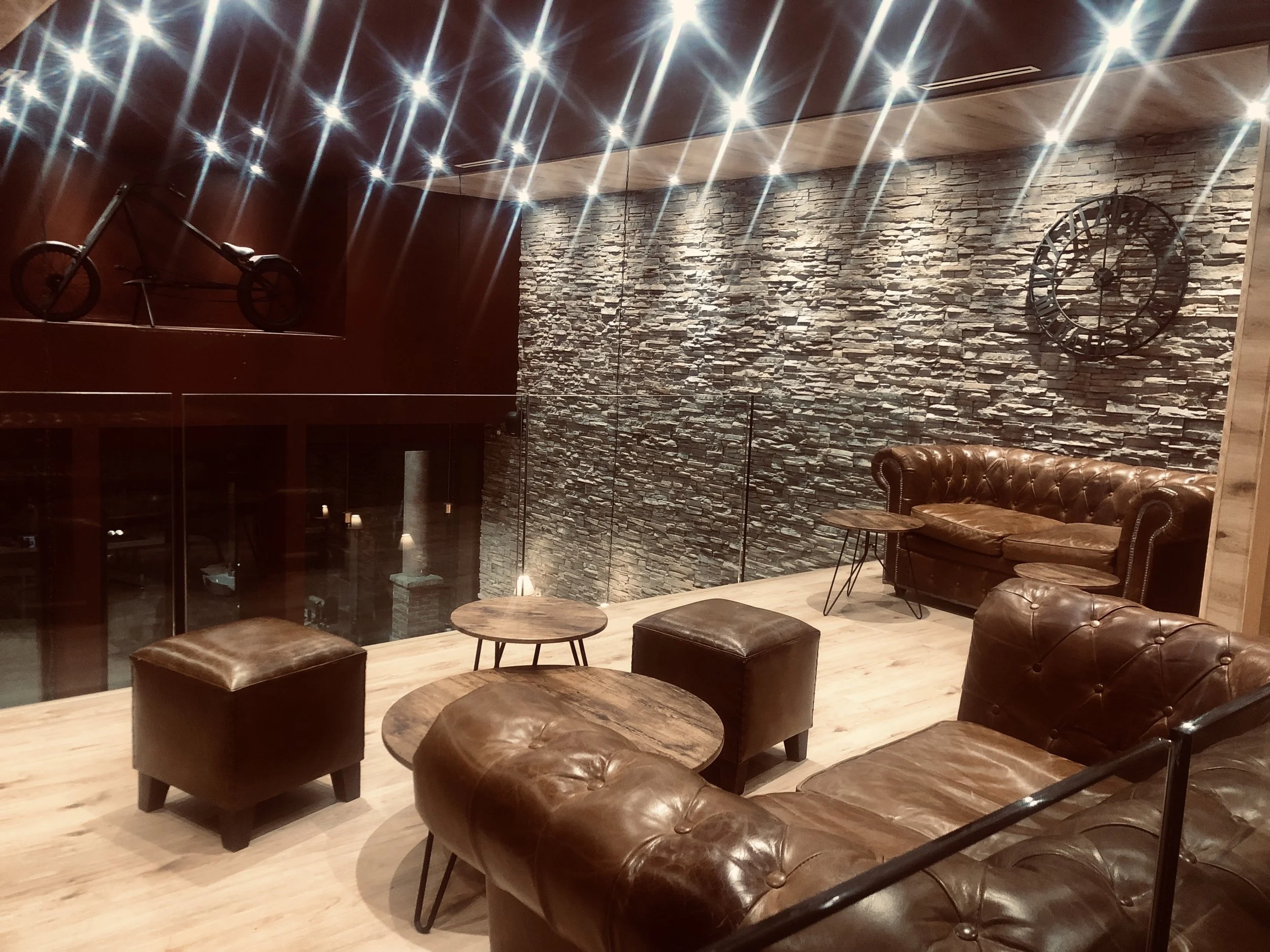 Salon intérieur avec canapés en cuir marron, tables en bois, mur en pierre, horloge murale, vélo suspendu au plafond, éclairage suspendu brillant. Barber shop salon de coiffure Marrakech