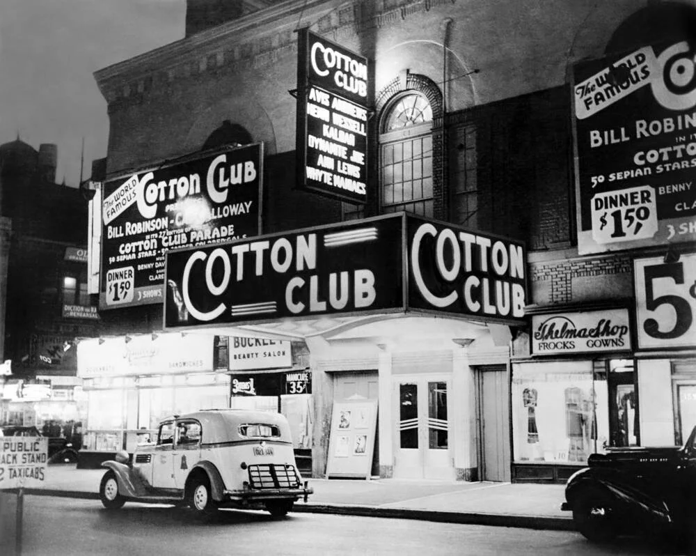 Une rue animée avec plusieurs enseignes lumineuses, notamment pour le Cotton Club et un salon de beauté, avec une voiture dans la rue.