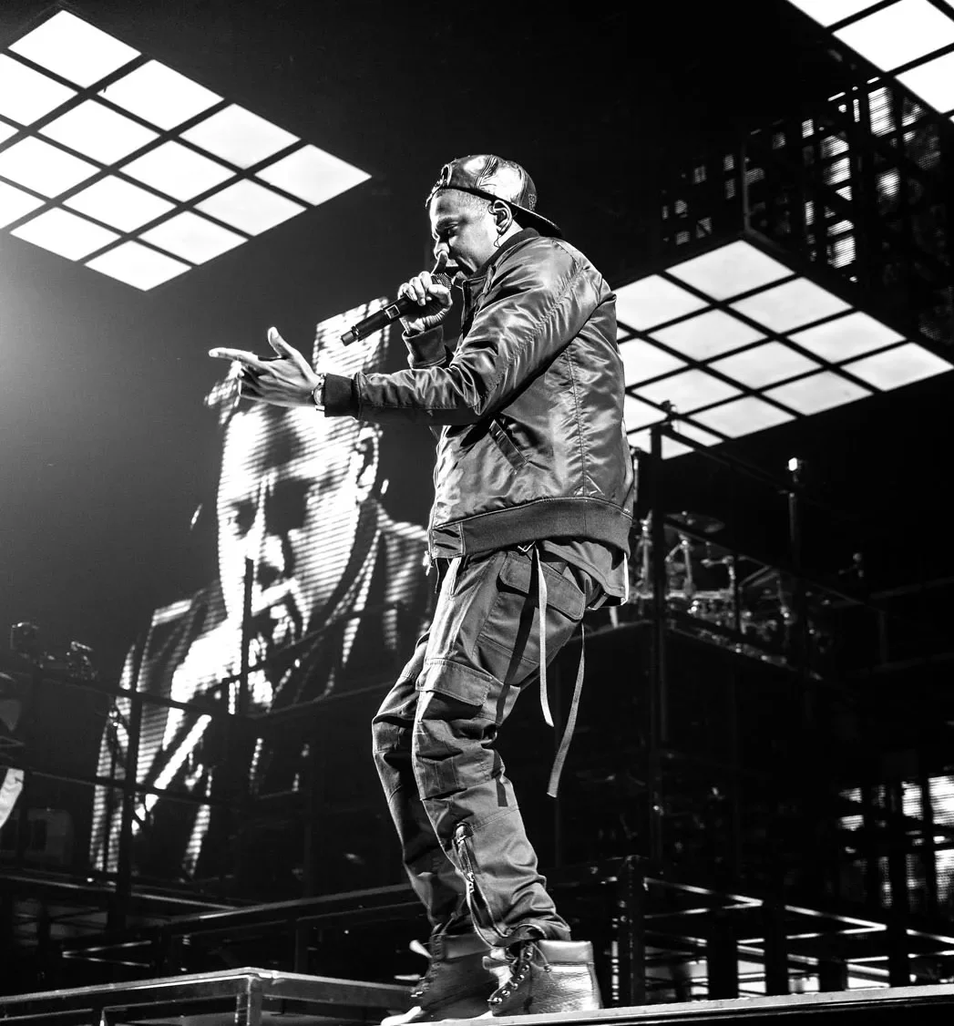 Un rappeur performe en noir et blanc sur scène, tenant un microphone, avec des écrans lumineux en arrière-plan.