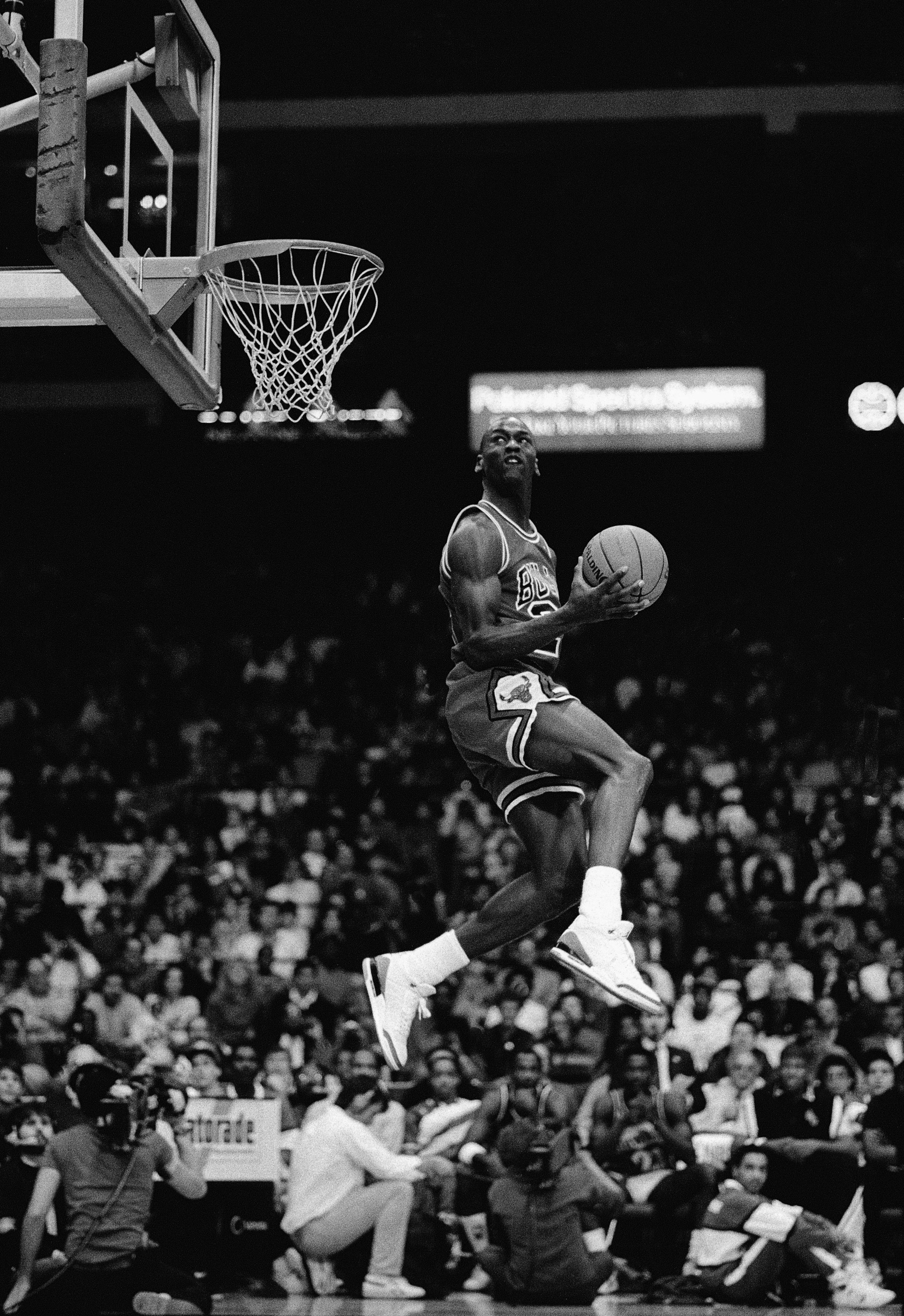 Un joueur de basketball en plein saut vers le panier, tenant le ballon, lors d'un match dans une salle bondée.