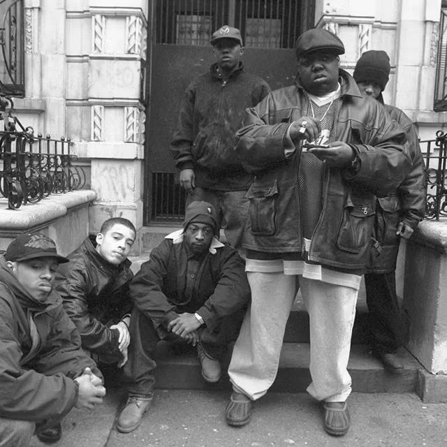 Groupe de six jeunes hommes posant devant un bâtiment en pierre, certains assis sur les marches et d'autres debout, en vêtements décontractés.