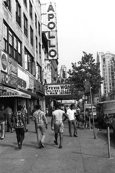 Rue animée avec des gens qui se promènent, des bâtiments avec des enseignes, y compris un théâtre Apollo, et des arbres en ville.