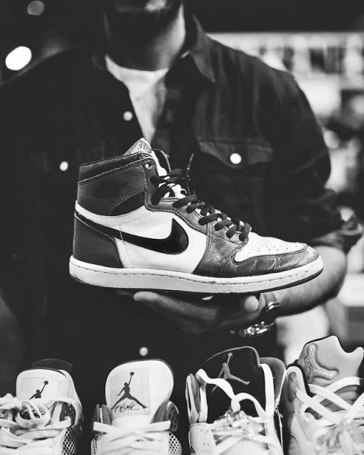 Une personne tient une chaussure Nike Air Jordan 1 à la main, avec plusieurs autres baskets en dessous. L'image est en noir et blanc.