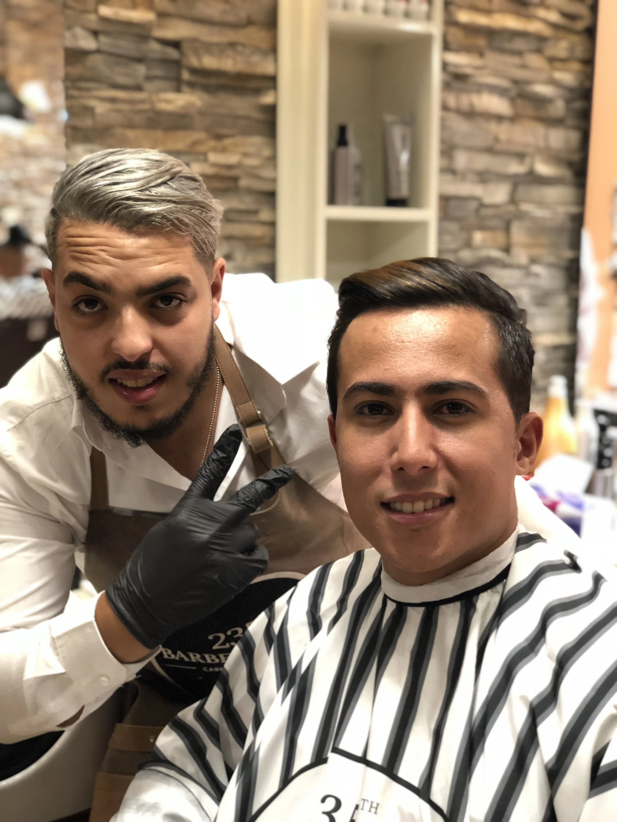 Deux jeunes hommes dans un salon de coiffure. L'un d'eux au premier plan porte une cape de coupe et sourit, l'autre derrière porte des gants noirs et fait un signe de paix à la caméra. Best barber shop salon de coiffure Marrakech