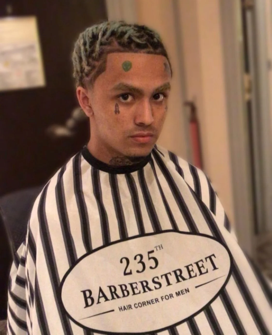 Jeune homme avec des tatouages, portant une cape de barbier avec le texte '235th Barberstreet Hair Corner For Men', dans un salon de coiffure. Best barber shop salon de coiffure Marrakech