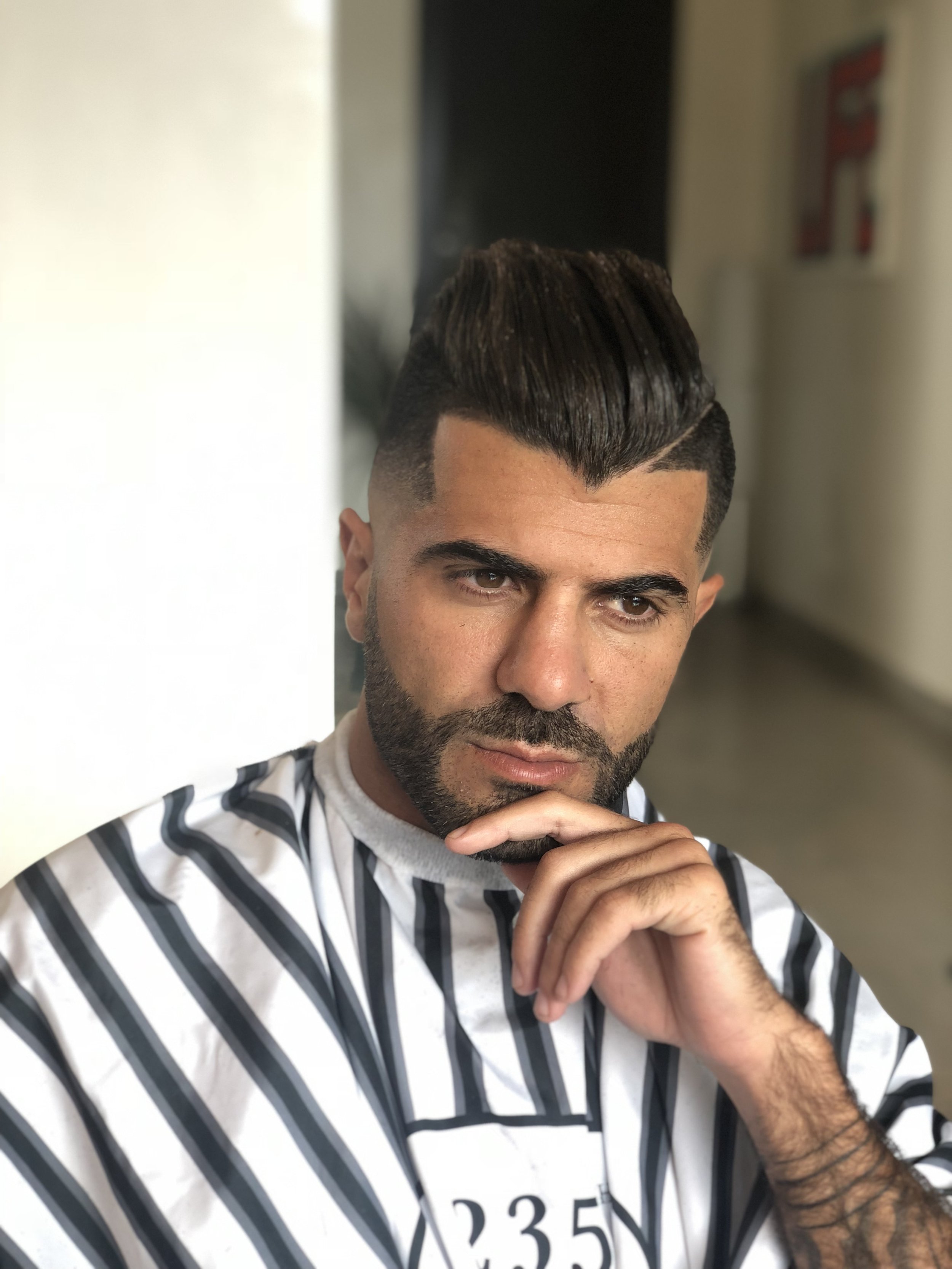 Un homme avec une coupe de cheveux moderne et une barbe, portant une veste rayée, assis dans un intérieur, du menton sur la main, regardant pensivement. Best barber shop salon de coiffure Marrakech