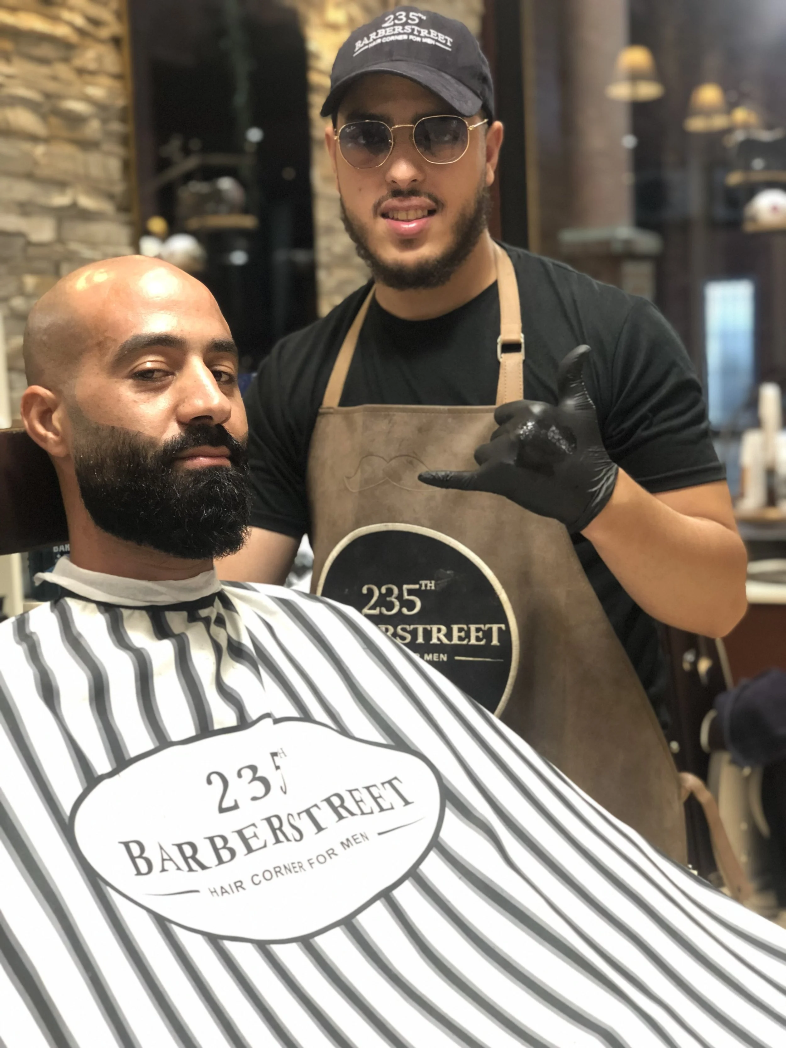 Un homme avec une barbe et une tête rasée reçoit une coupe de cheveux dans un salon de coiffure appelé '235th BarberStreet'. Un autre homme, portant un tablier avec le même logo, se tient derrière lui, faisant un signe de la main. Le décor du salon inclut un mur en pierre et des éléments en bois.