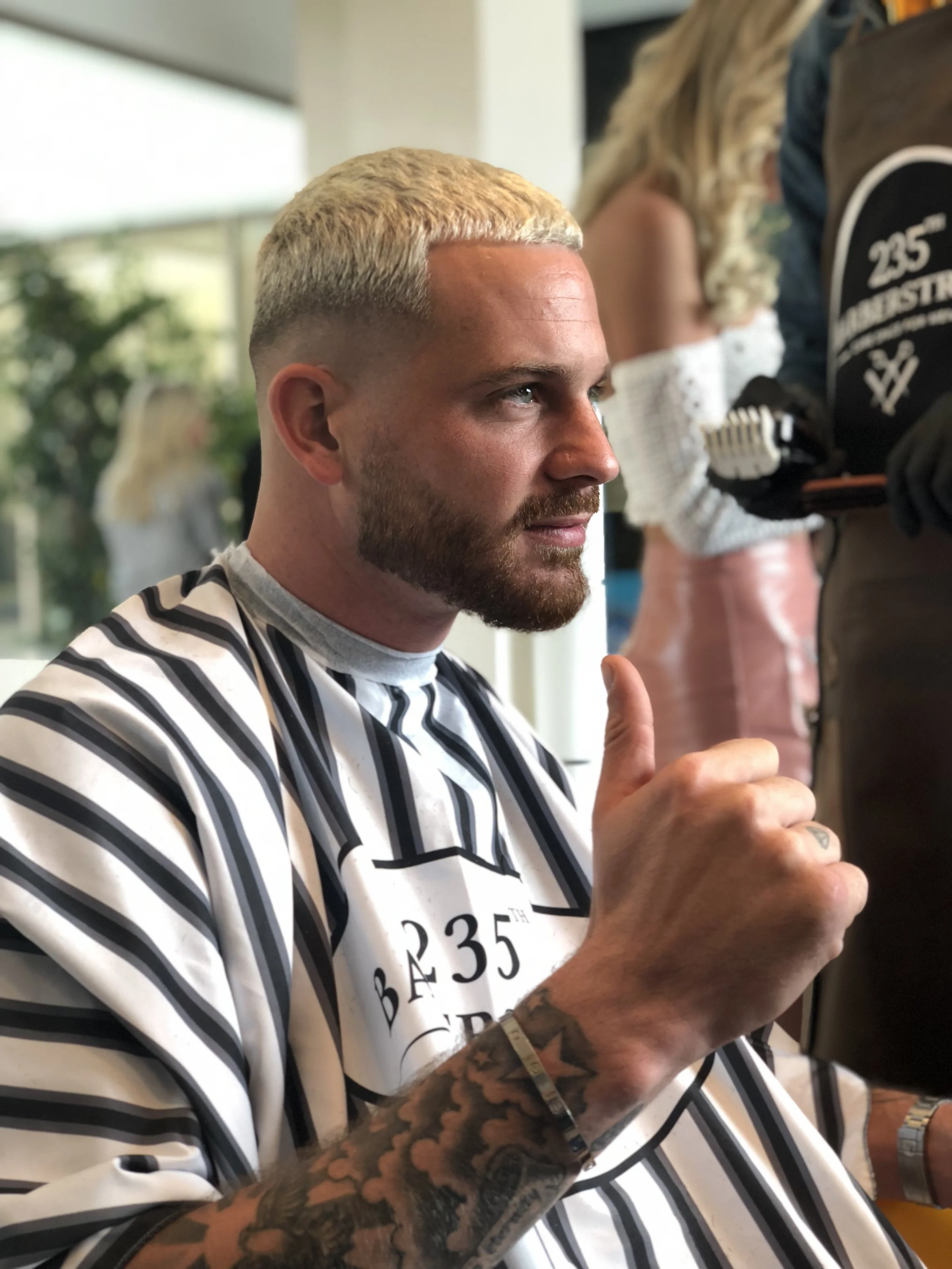 Un homme avec une barbe, aux cheveux blonds, portant une cape de coupe de cheveux à rayures noires et blanches, faisant un geste de pouce levé. Best barber shop salon de coiffure Marrakech