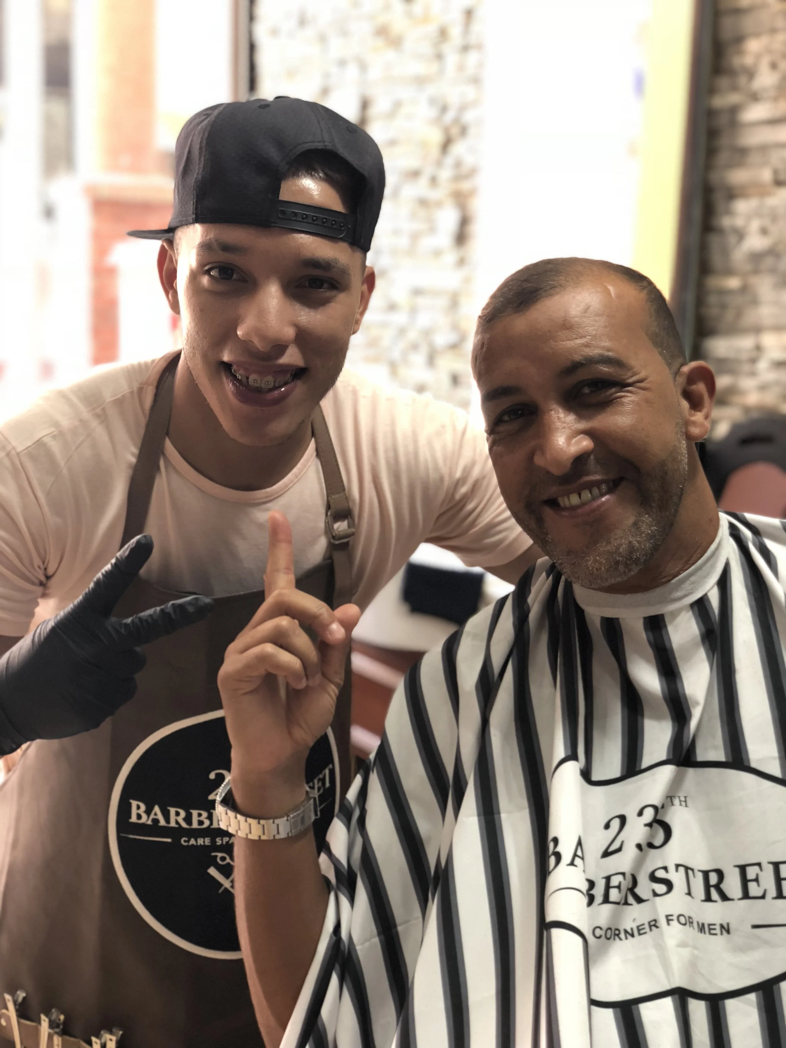 Deux hommes posent pour une photo, l'un d'eux porte une cape de coiffure avec l'inscription 'Barberstreet' et l'autre fait un geste de doigt levé, tous deux souriants. Best barber shop salon de coiffure Marrakech
