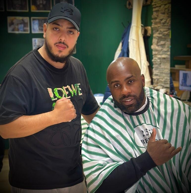 Deux hommes posent dans un salon de coiffure, l'un portant une cape à rayures et faisant un signe de la main, l'autre portant une casquette noire. Best barber shop salon de coiffure Marrakech