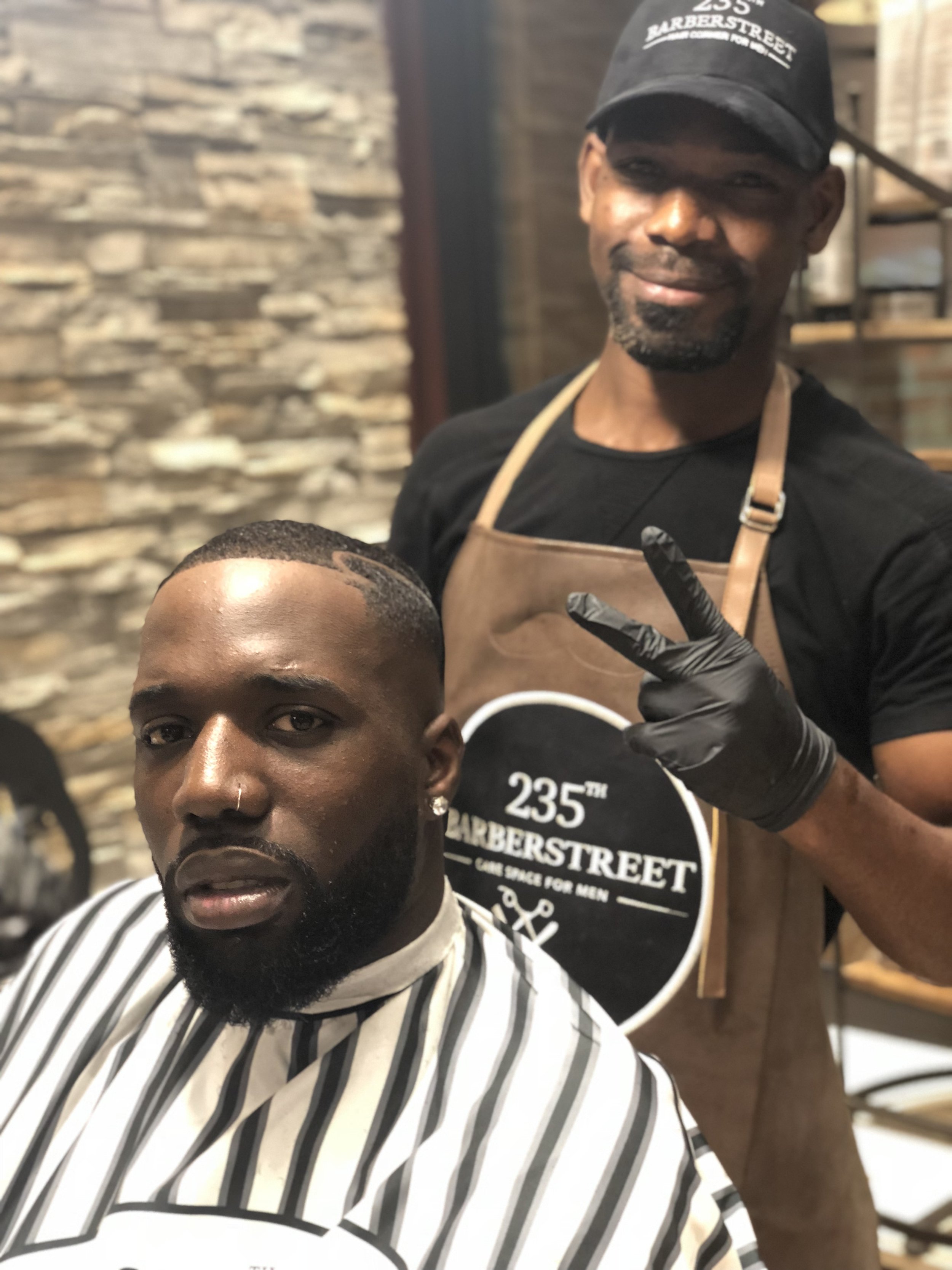 Un homme dans un fauteuil de salon de coiffure portant une cape rayée, recevant une coupe de cheveux par un barbier noir portant un tablier en cuir et un gant noir, dans un salon avec un mur en pierre en arrière-plan. Best barber shop 