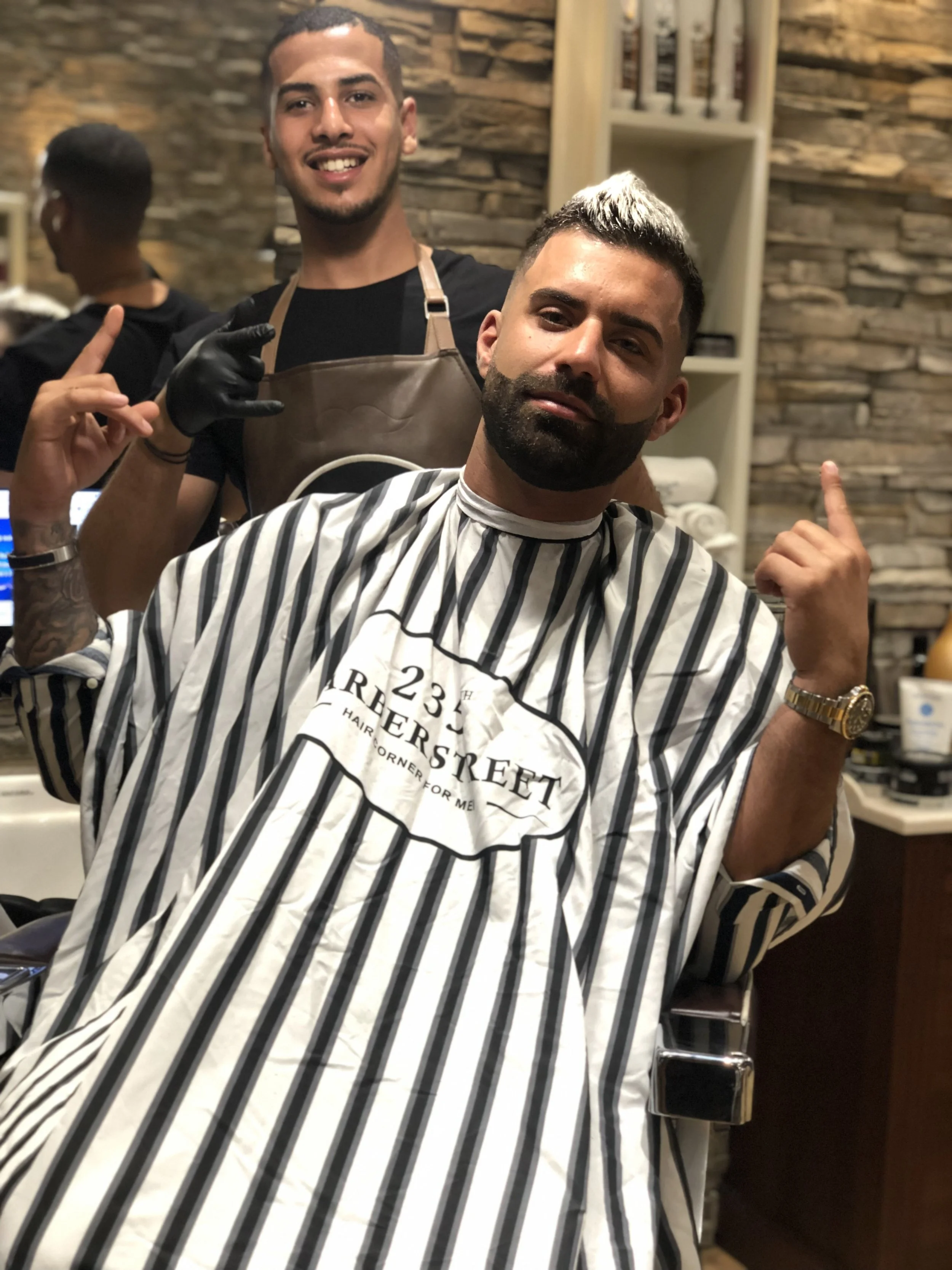 Une personne est assise dans un fauteuil de salon de coiffure, portant une cape à rayures noires et blanches. Deux autres personnes se tiennent derrière, l'une en t-shirt noir et la seconde un tablier de coiffeur, tous dans un décor avec mur en pierre et étagères.