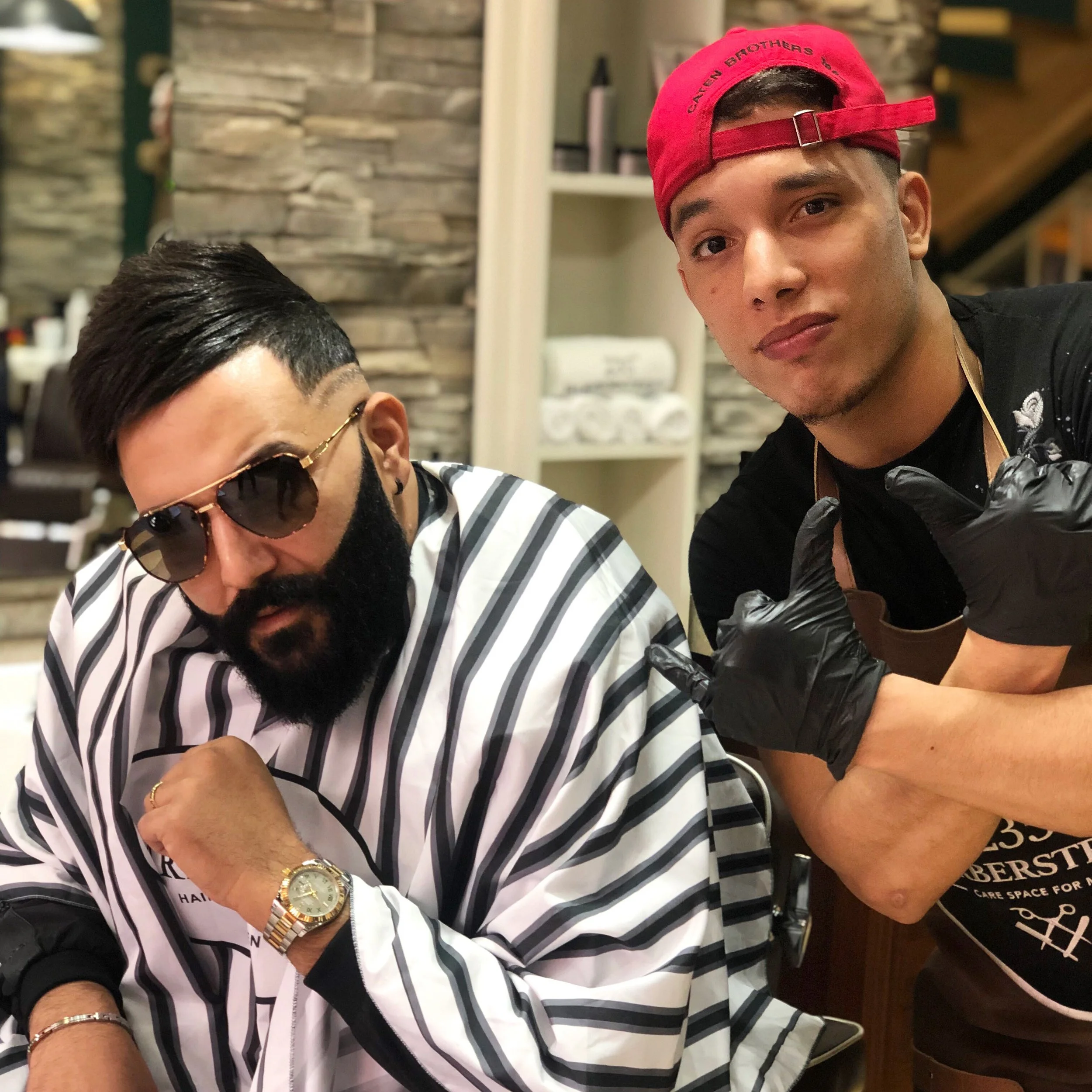 Deux hommes posant dans un salon de coiffure ou un barbershop. L'un porte des lunettes de soleil et une chemise rayée, l'autre porte un chapeau rouge et un tablier de barbier, faisant un geste de bras croisés. Best barber shop salon de coiffure 
