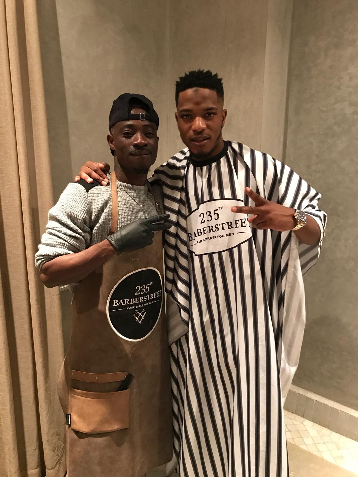 Deux hommes posent ensemble, un porte une robe de barbier et l'autre porte une robe de barbier avec des rayures, dans un intérieur. Best barber shop salon de coiffure Marrakech