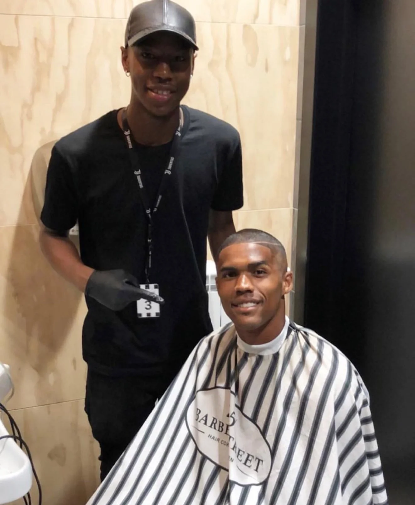 Un homme qui reçoit une coupe de cheveux dans un salon, assis dans une cape de barbier avec le logo « BARBER STREET ». Un autre homme, portant un t-shirt noir, une casquette, et une étiquette d'identification, se tient à côté. Best barber shop maroc