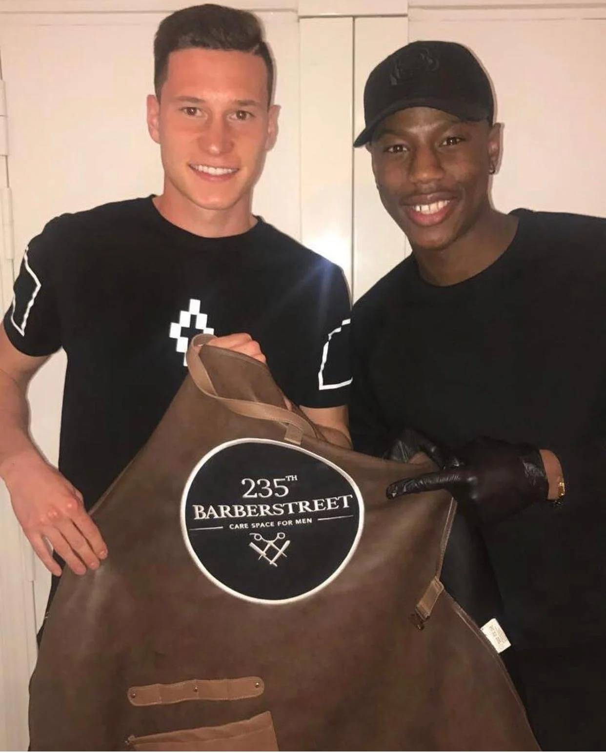 Deux jeunes hommes posent pour une photo, l'un porte un t-shirt noir et l'autre porte un noir cap, ils tiennent un grand tablier de barbiers avec une étiquette indiquant 235th Barberstreet. Best barber shop salon de coiffure Marrakech