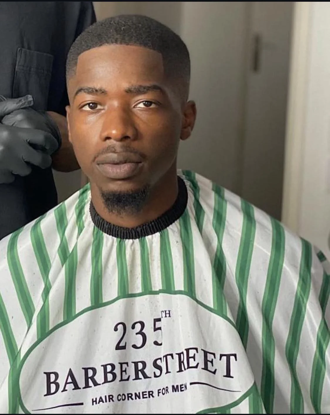Jeune homme assis en salon de coiffure, portant une cape rayée vert et blanc avec inscription « 235th Barber Street Hair Corner for Men », après avoir été rasé, avec un fond simple d'une porte et un mur. Best barber shop salon de coiffure Marrakech