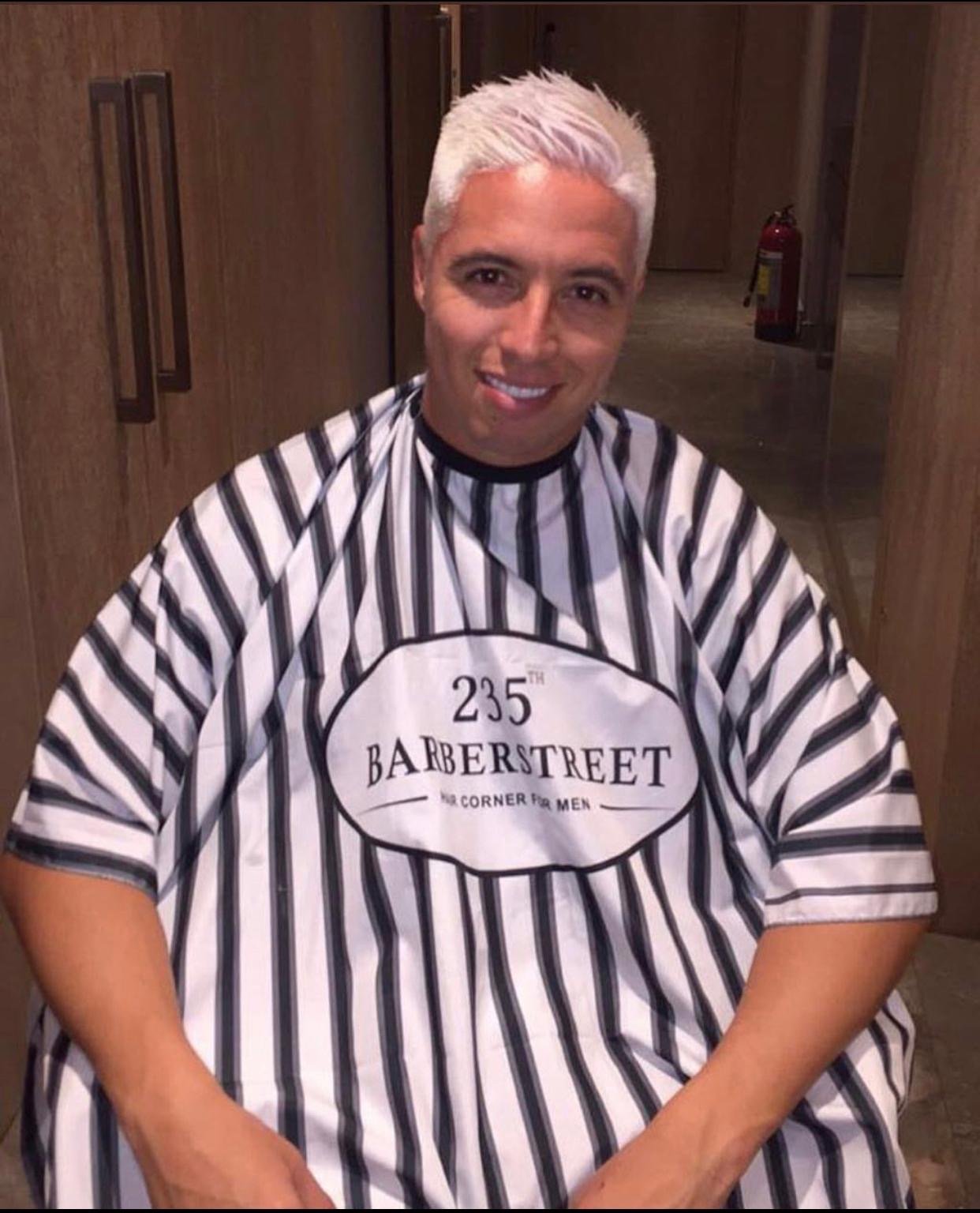 Homme avec cheveux blonds platinés en coiffure courte, portant une cape de salon de coiffure rayée noire et blanche, sur un fond d'intérieur en bois. Best barber shop salon de coiffure Marrakech