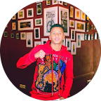 Jeune homme souriant avec une veste colorée, tenant un appareil photo, dans un décor artistique avec des œuvres d'art sur le mur.