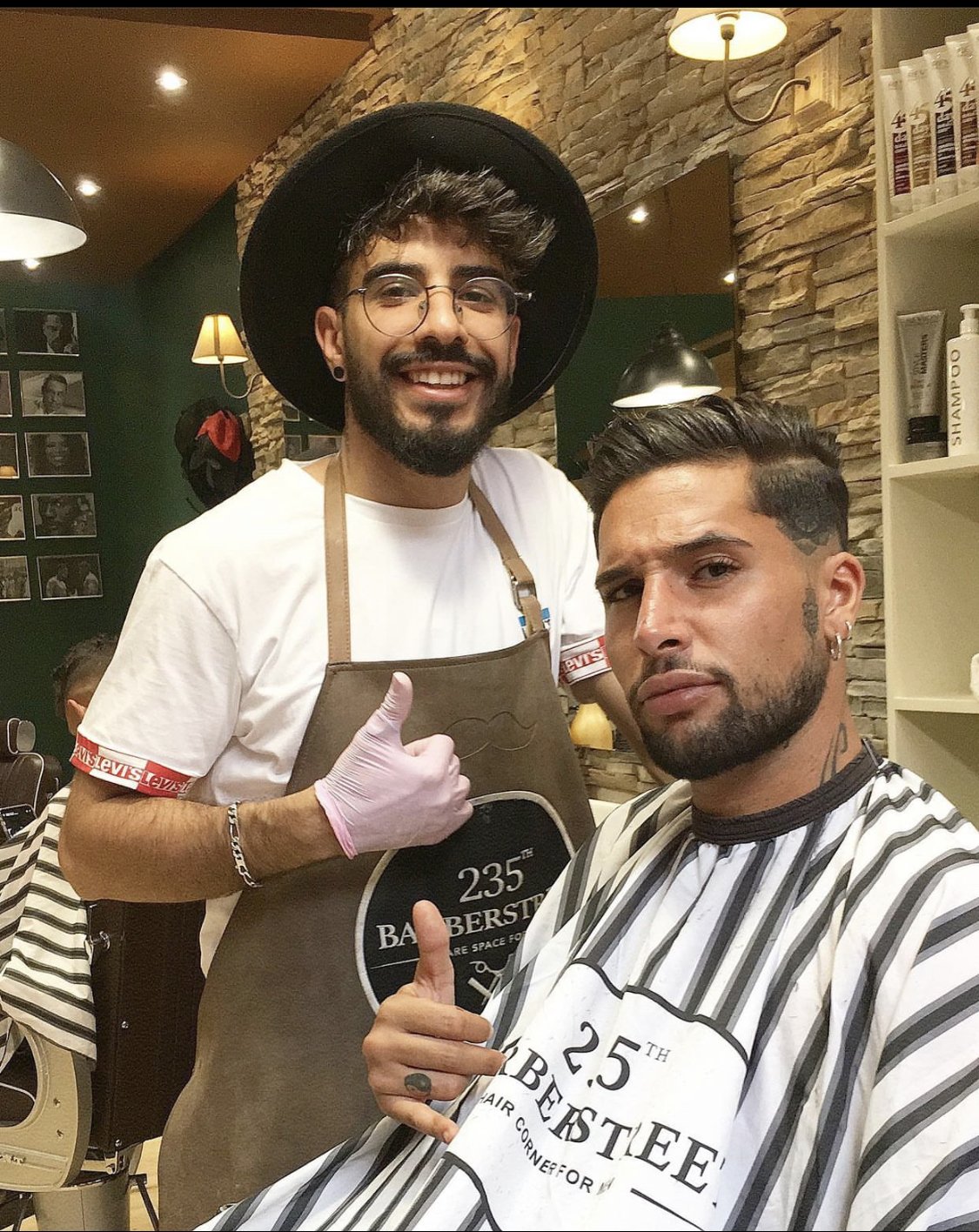 Deux hommes dans un salon de coiffure. L'un d'eux est assis, portant une cape de coiffeur avec l'inscription '25th BARBERSTEE'. L'autre, debout, porte un tablier de barbier avec la même inscription et un chapeau noir, lui faisant un signe de pouce levé. La décoration du salon inclut un mur en pierre, un miroir, et des photos en noir et blanc au mur.