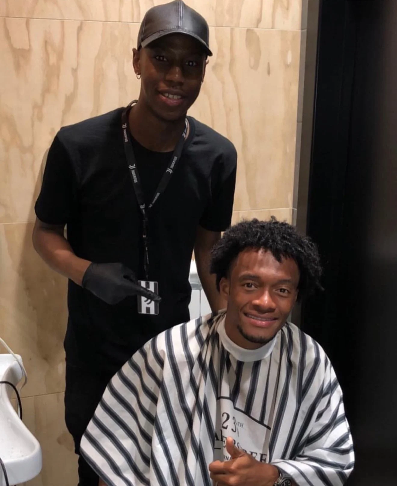 Deux hommes dans un salon de coiffure, un assis avec une cape à rayures et l'autre debout, portant un casque en cuir et un badge. Best barber shop salon de coiffure Marrakech