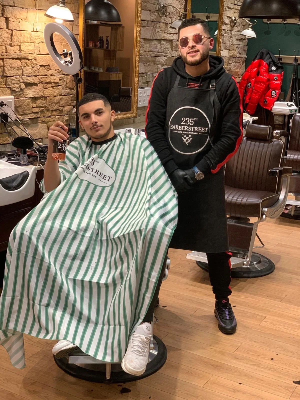 Un homme est assis dans un fauteuil de barbier avec une cape à rayures vertes et blanches, tenant une bouteille. Un autre homme, barbier, se tient à côté, portant un tablier noir et des lunettes de soleil, dans un salon de coiffure avec un décor en pierre et un grand miroir.