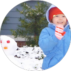 Un enfant souriant en tenue d'hiver avec un bonnet de Noël, dehors avec de la neige et un arbre en arrière-plan.