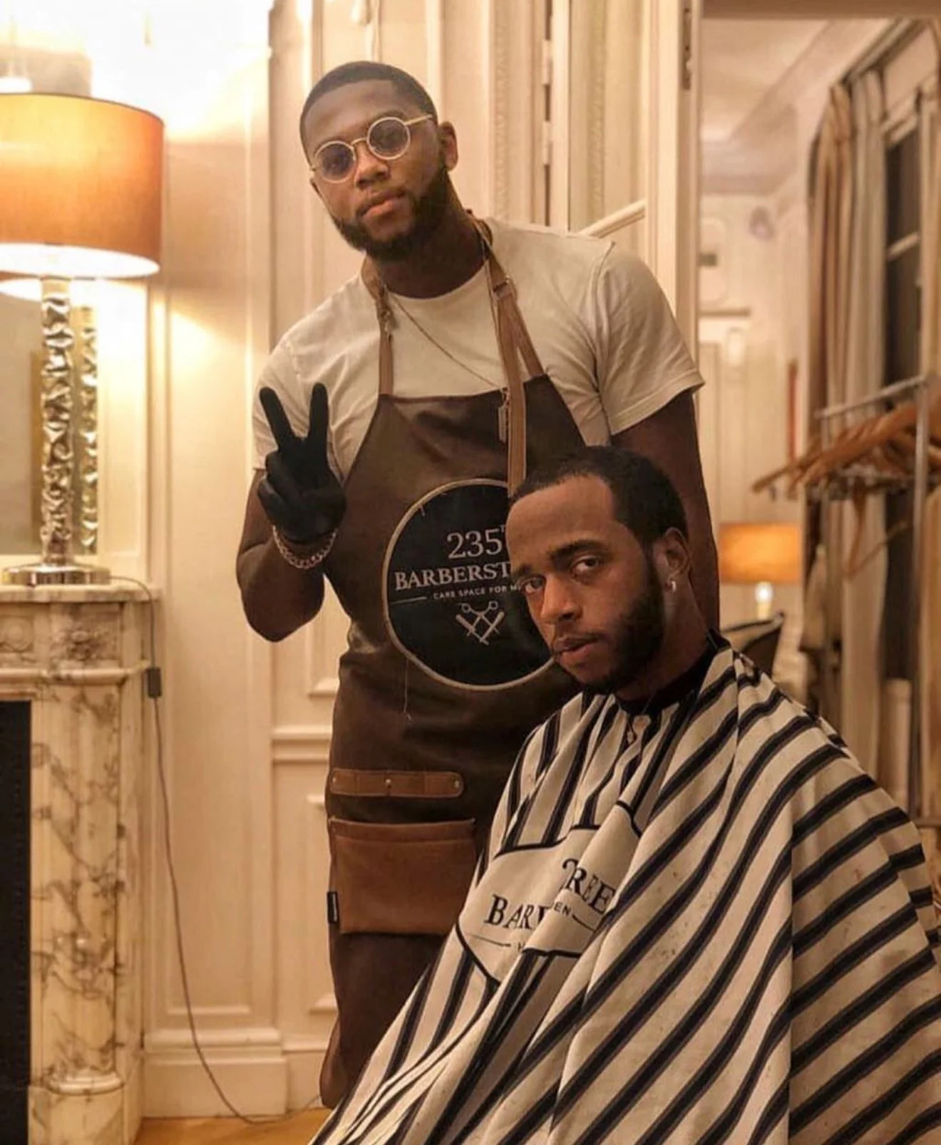 Deux hommes dans un salon de coiffure. L'un porte une cape de barbier, l'autre un tablier de barbier. L'homme derrière porte des lunettes rondes et fait un signe de paix avec la main. Best barber shop salon de coiffure Marrakech