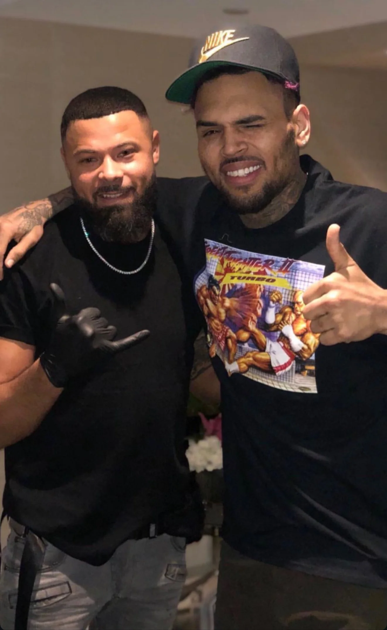 Deux hommes posent ensemble, l'un portant une casquette Nike et un t-shirt avec une scène de combat de Street Fighter, l'autre portant un t-shirt noir et faisant un signe de la main. Ils semblent heureux, dans un intérieur. Best barber shop 