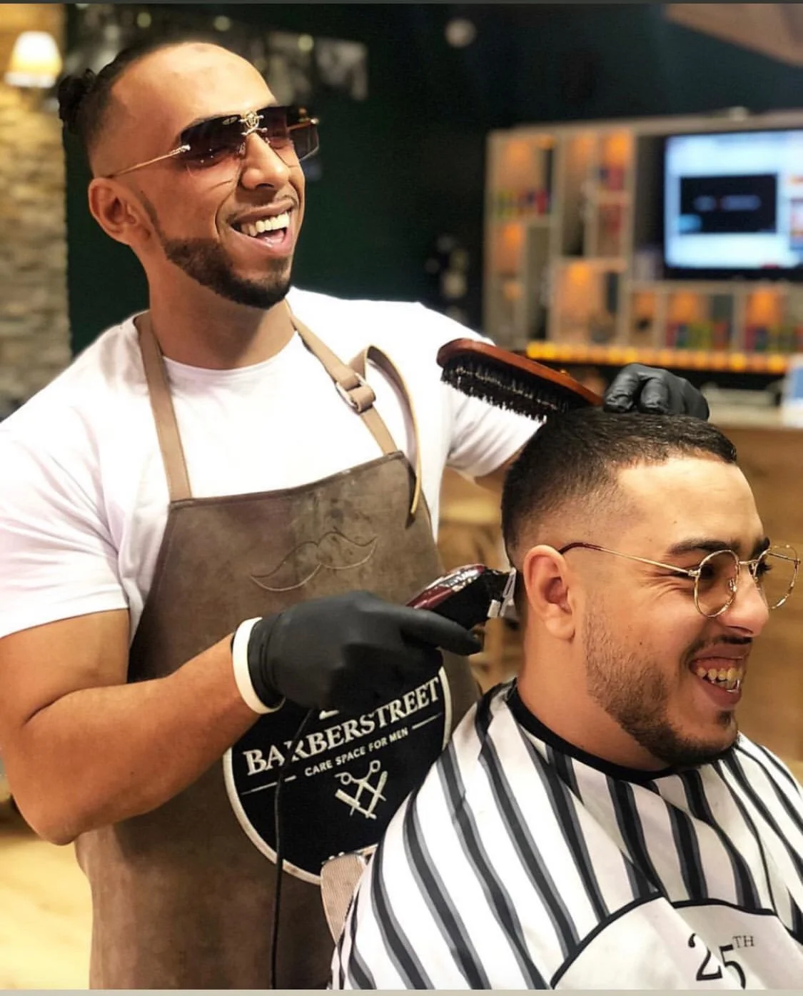 Un homme en train de barber un autre homme dans un salon de coiffure, tous deux souriants, portant des lunettes et un tablier, avec des outils de coiffure à la main. Best barber shop salon de coiffure Marrakech