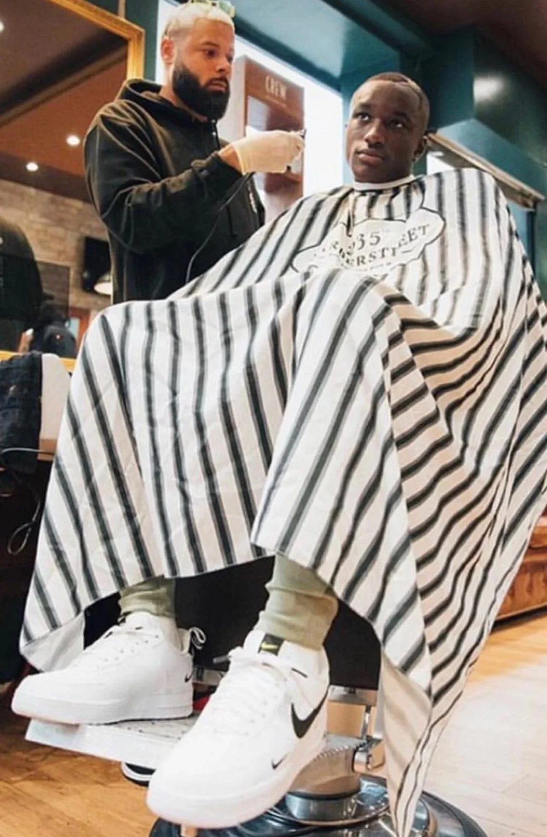 Un homme à une coupe de cheveux très courte ou rasée reçoit une coupe de cheveux dans un salon de coiffure, avec un peignoir à rayures noirs et blanches. Un coiffeur porte des gants et tient un outil de coupe, observant l'homme. La scène se déroule dans un intérieur de salon avec un mur de briques et des lampes.