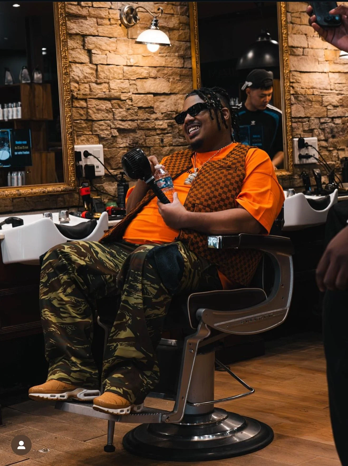 Un homme assis dans un fauteuil de barberie, portant des vêtements de style urbain, tenant un microphone, souriant et portant des lunettes de soleil. Il y a un grand miroir derrière lui, une lampe suspendue au plafond, et une personne en arrière-plan dans un environnement de salon de coiffure.