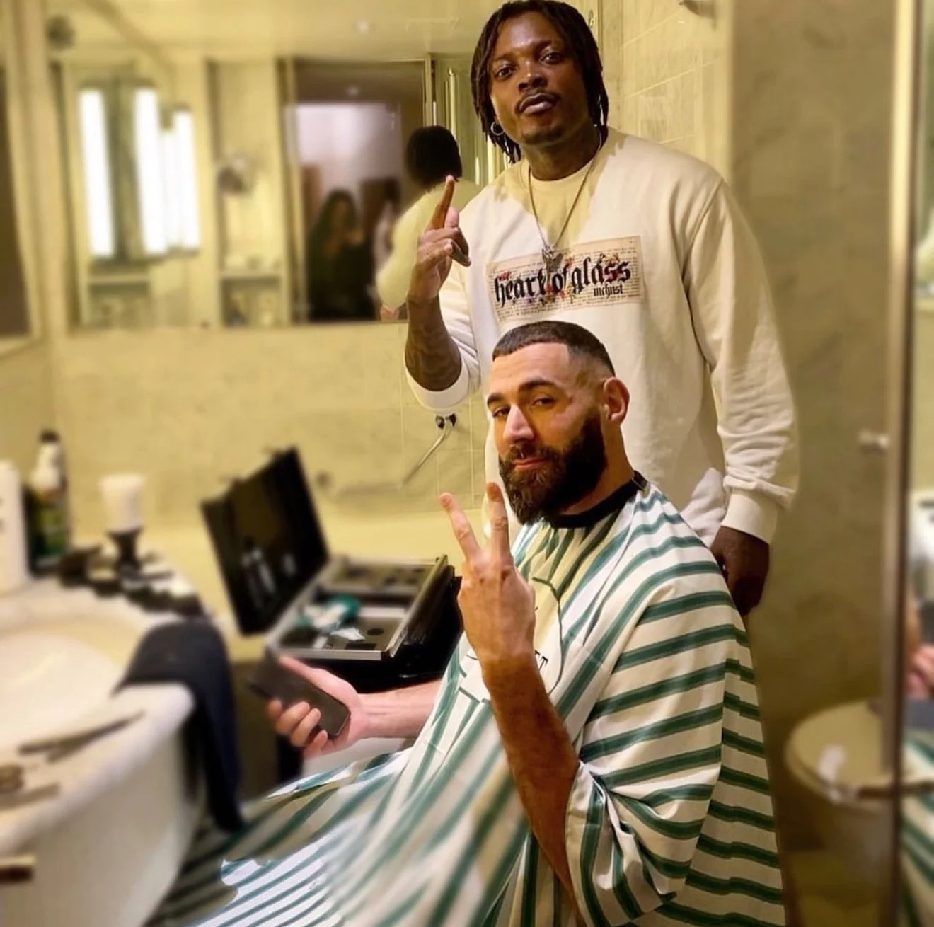 Un homme avec une barbe portant une cape de barbier et tenant un téléphone, faisant un signe de paix avec ses doigts, assis dans un salon de coiffure. Un autre homme derrière lui, avec des dreadlocks, portant un sweat-shirt blanc avec un texte en anglais, fait un signe de paix avec sa main. Deux autres personnes peuvent être vue dans un miroir en arrière-plan.